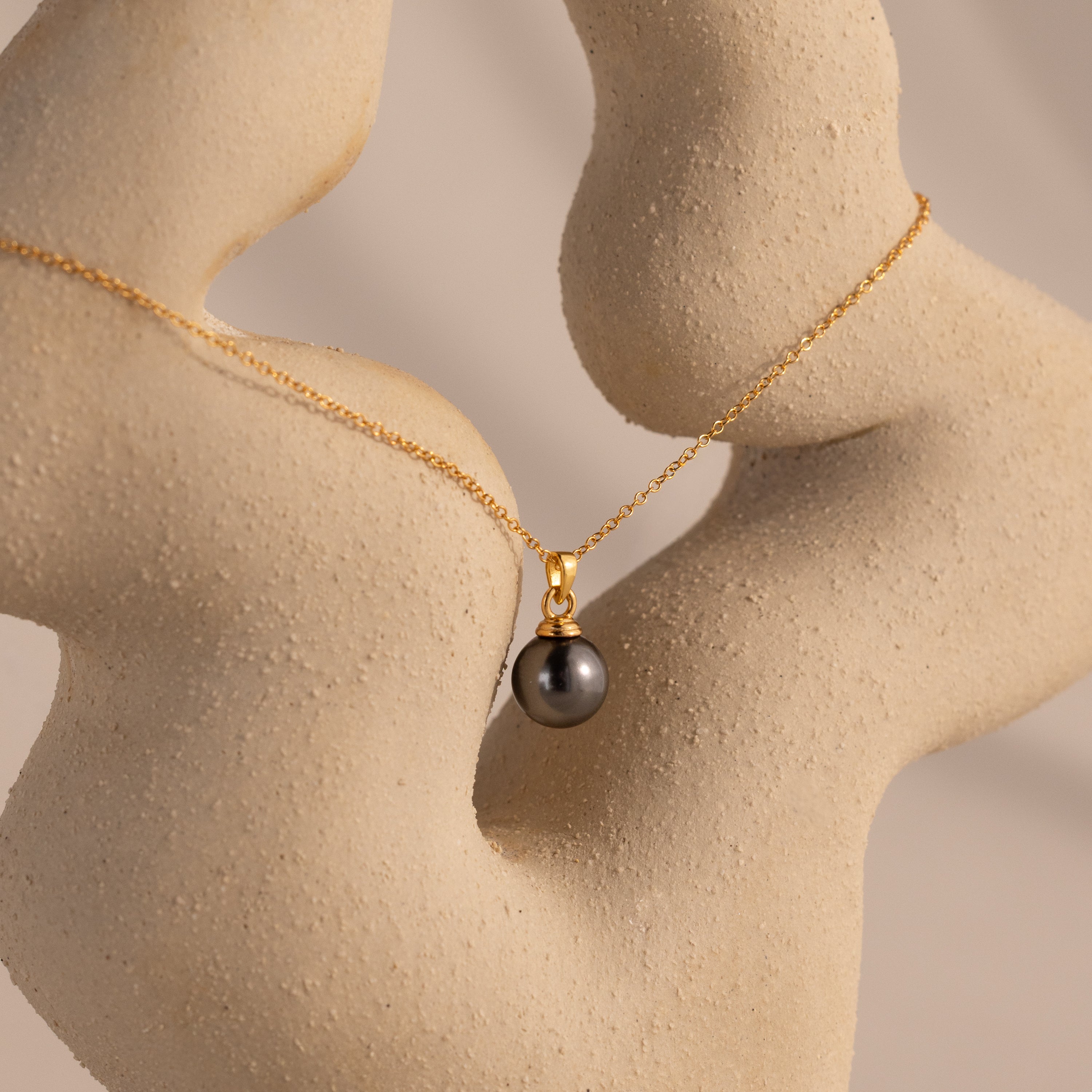 The Midnight Pearl Necklace in 18K Gold, featuring a black pearl pendant, is elegantly displayed draped over a textured abstract beige sculpture—a stunning jewelry piece.