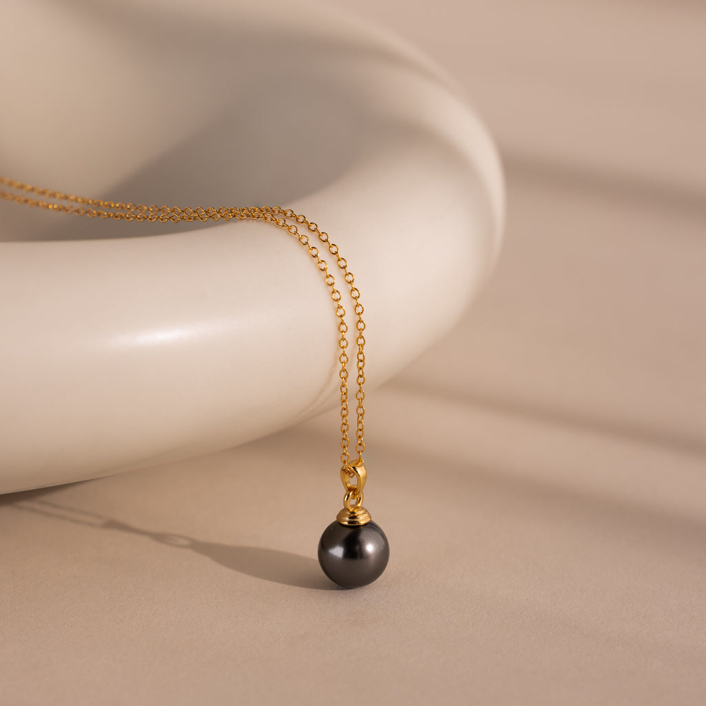 The Midnight Pearl Necklace in 18K Gold features a gold chain with a black pearl pendant, beautifully displayed on a smooth, cream-colored ceramic surface.