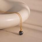 The Midnight Pearl Necklace in 18K Gold features a gold chain with a black pearl pendant, beautifully displayed on a smooth, cream-colored ceramic surface.