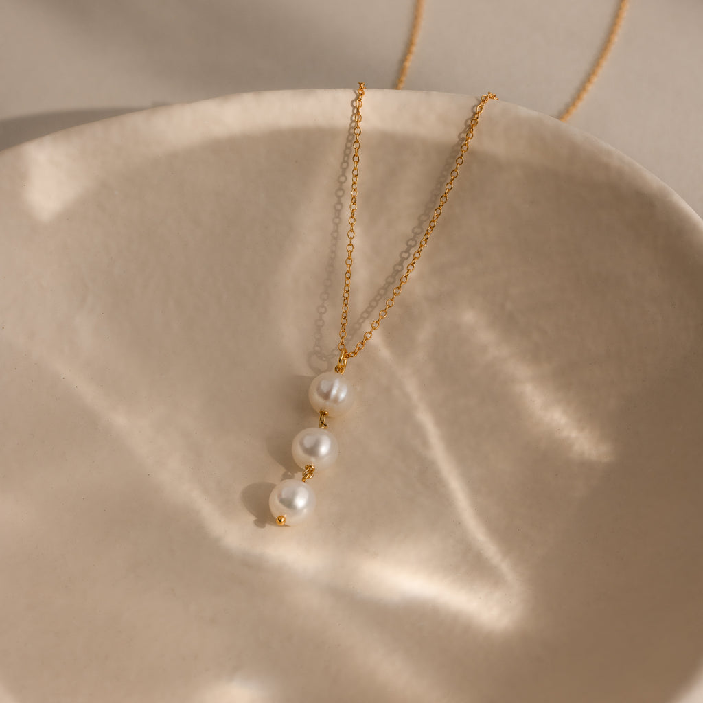Experience elegant jewelry at its finest with the Pearl Cascade Necklace in 18K Gold, featuring three white pearls beautifully displayed on a textured cream-hued ceramic dish—perfect for everyday wear.