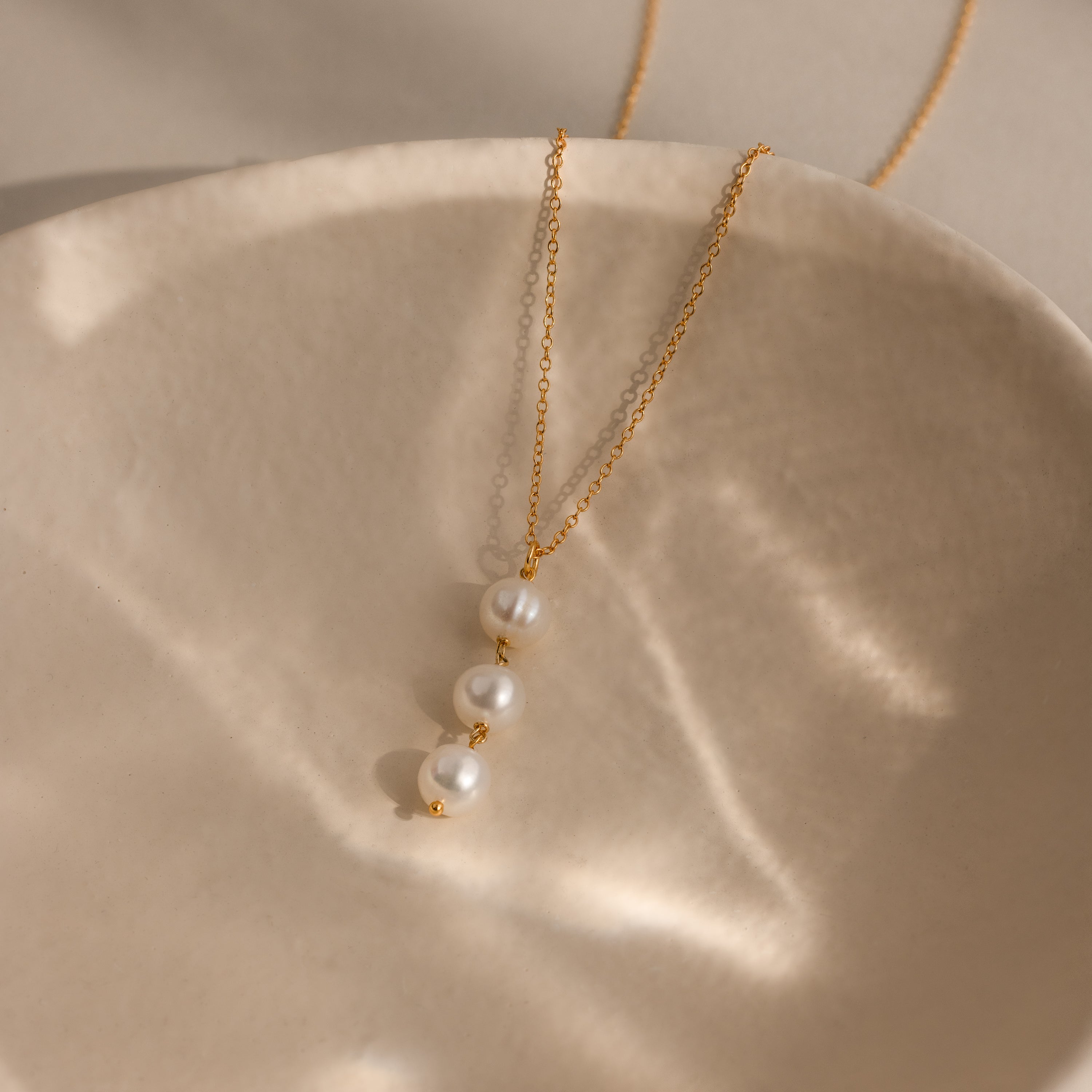 Experience elegant jewelry at its finest with the Pearl Cascade Necklace in 18K Gold, featuring three white pearls beautifully displayed on a textured cream-hued ceramic dish—perfect for everyday wear.