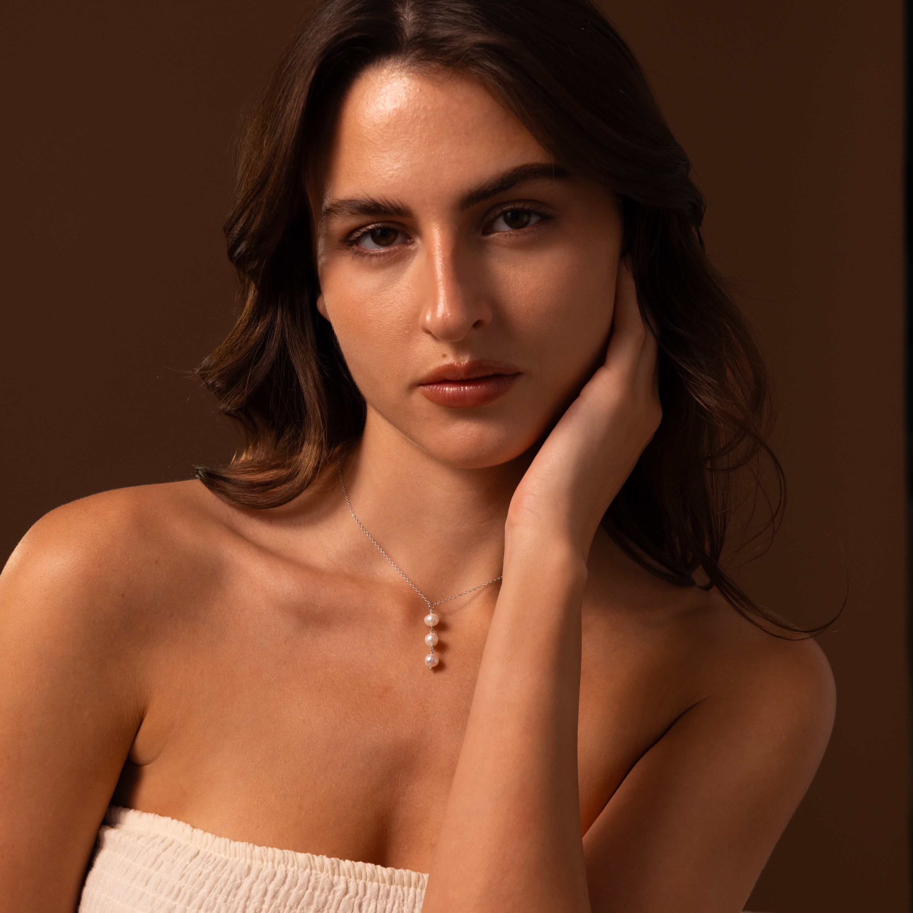 A woman with long brown hair rests her head on her hand, wearing a strapless top and the Pearl Cascade Necklace in Sterling Silver, an elegant accessory featuring luminous pearls.