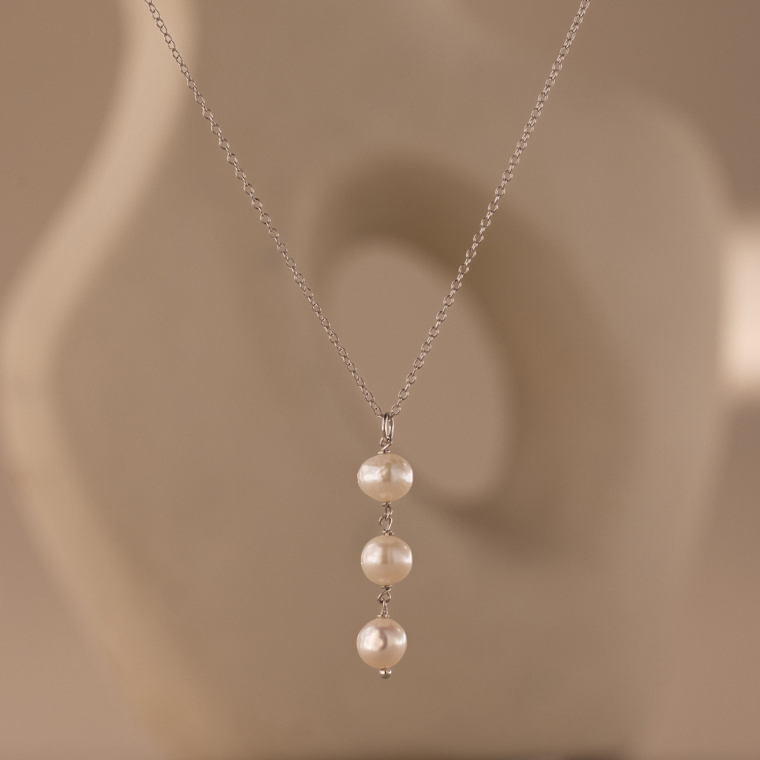 The Pearl Cascade Necklace in Sterling Silver features three luminous pearls hanging vertically on a silver chain, set against a soft beige abstract background.