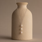 The Pearl Cascade Necklace in Sterling Silver, featuring luminous pearls on a sleek silver chain, is elegantly draped over a textured beige vase.
