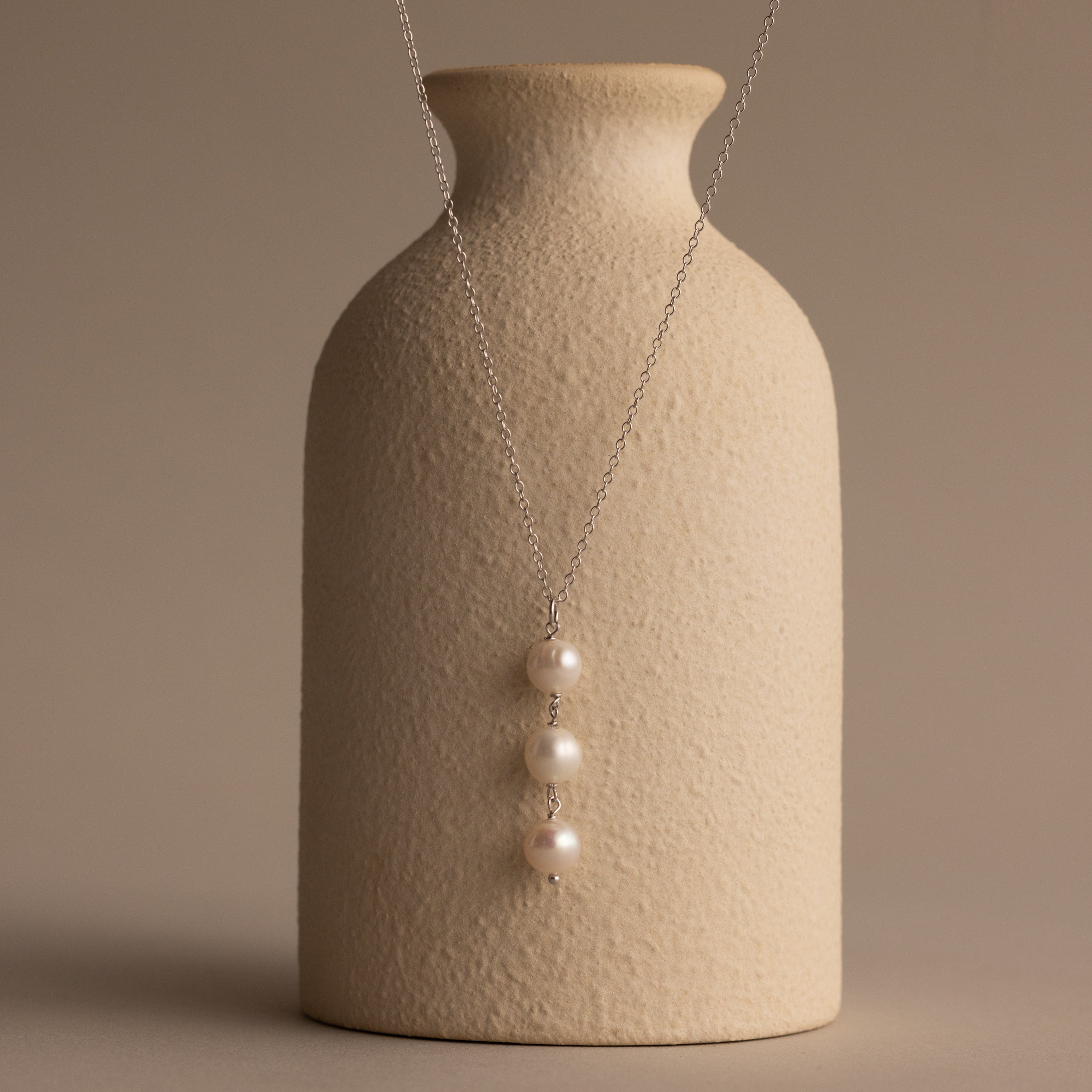 The Pearl Cascade Necklace in Sterling Silver, featuring luminous pearls on a sleek silver chain, is elegantly draped over a textured beige vase.