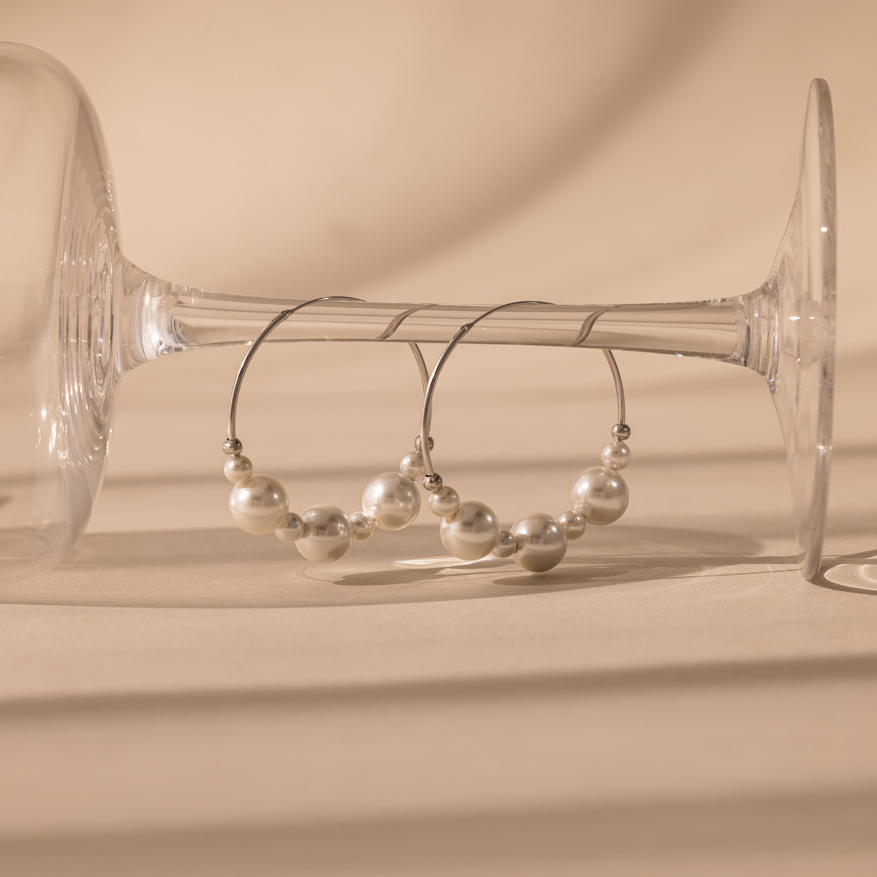 The Statement Pearl Hoops in Sterling Silver hang elegantly on a wine glass stem against a beige background, highlighting the refined allure of luxury jewelry.