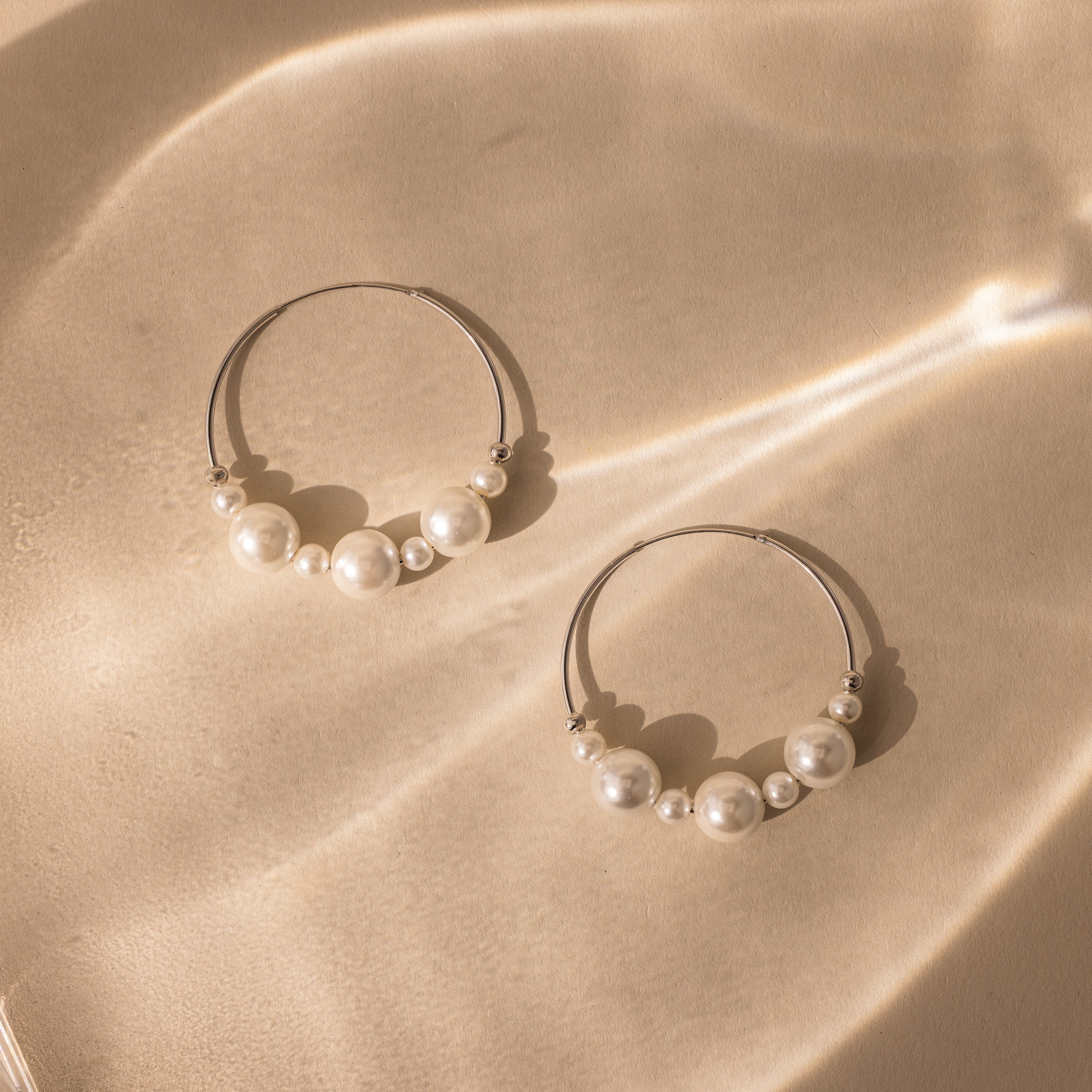 Statement Pearl Hoops in Sterling Silver with white pearls rest on a beige surface, illuminated by soft, wavy light—an elegant showcase of luxurious jewelry.