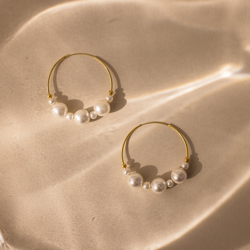 Statement Pearl Hoops featuring an open hoop design with luminous white pearls, shown on a beige surface highlighted by soft, wavy light reflections.