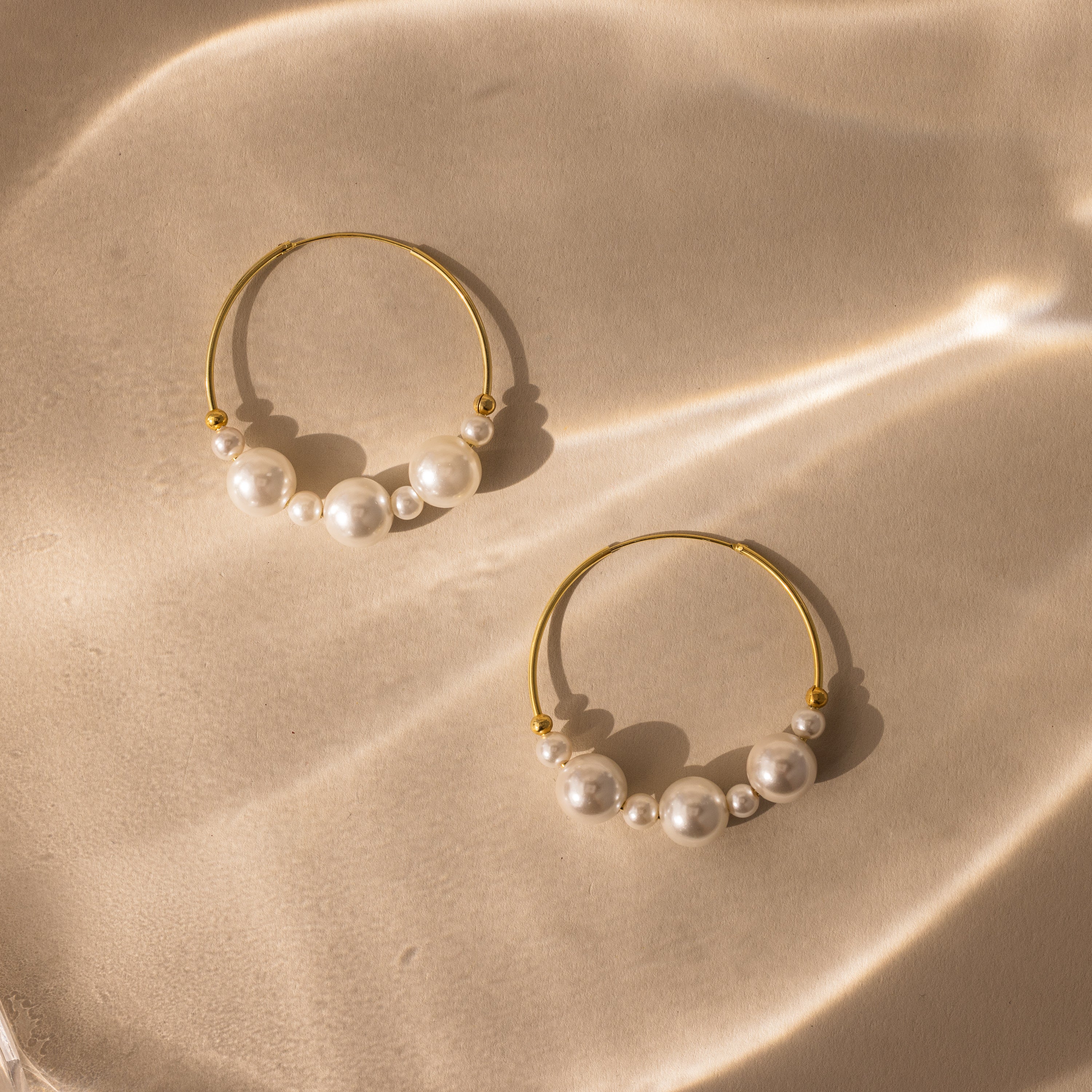 Statement Pearl Hoops featuring an open hoop design with luminous white pearls, shown on a beige surface highlighted by soft, wavy light reflections.