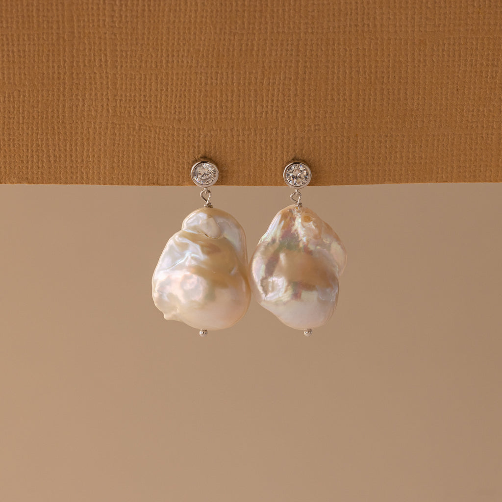 The Diamond Baroque Pearl Studs in Sterling Silver are displayed against a textured beige background, showcasing their organic pearl design with round diamond studs and baroque pearls.