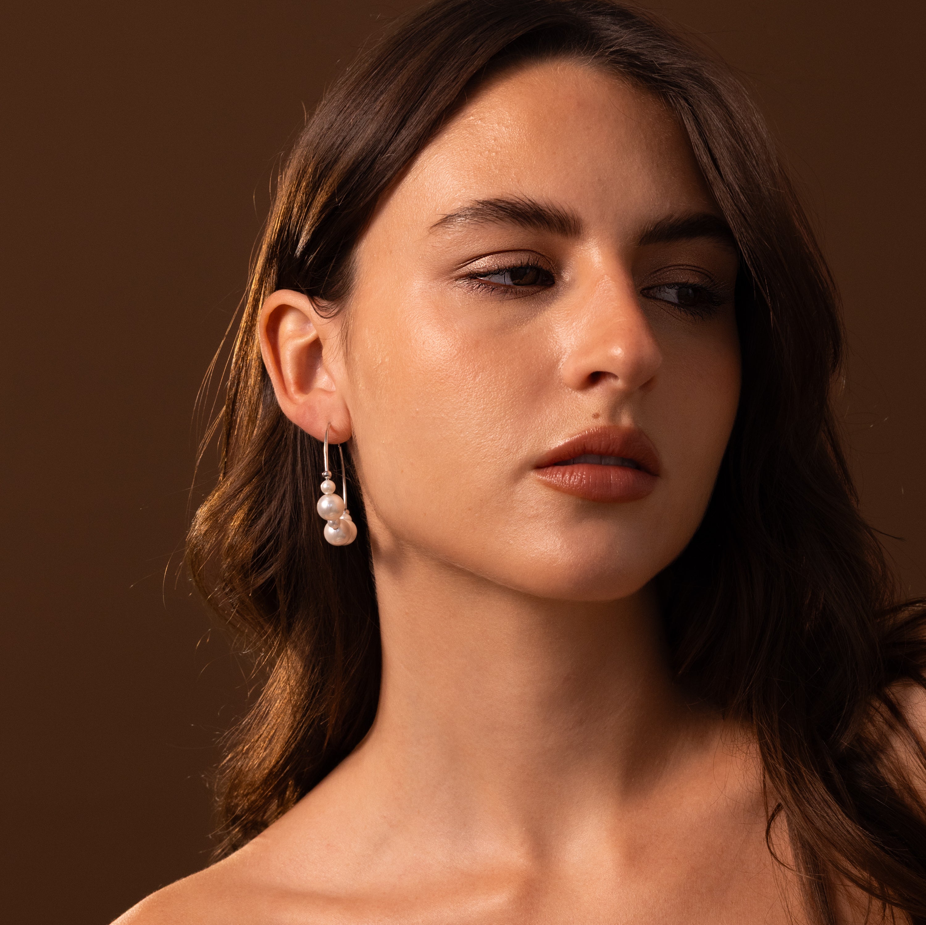 A woman with long brown hair wears Statement Pearl Hoops in Sterling Silver, gazing to the side against a brown background.