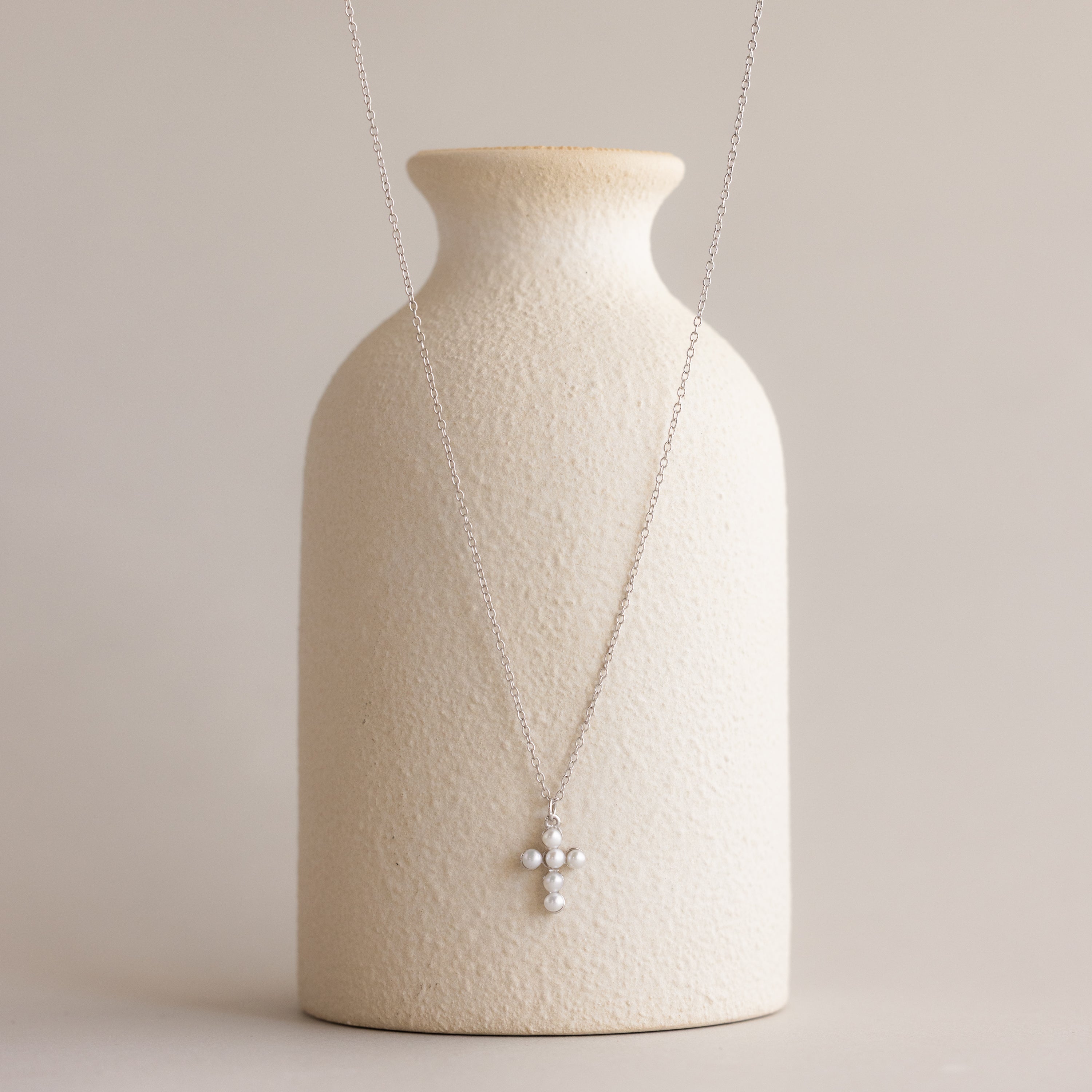 Pearl Cross Necklace in Silver showcased with luminous pearls on a textured cream vase.