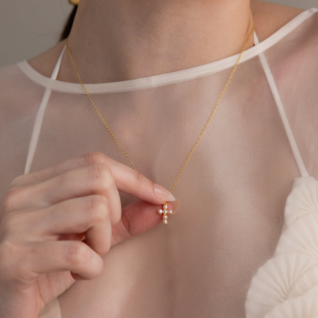 A woman wearing a sheer white top holds the Pearl Cross Necklace, featuring a gold chain with a delicate pearl and crystal cross charm pendant.