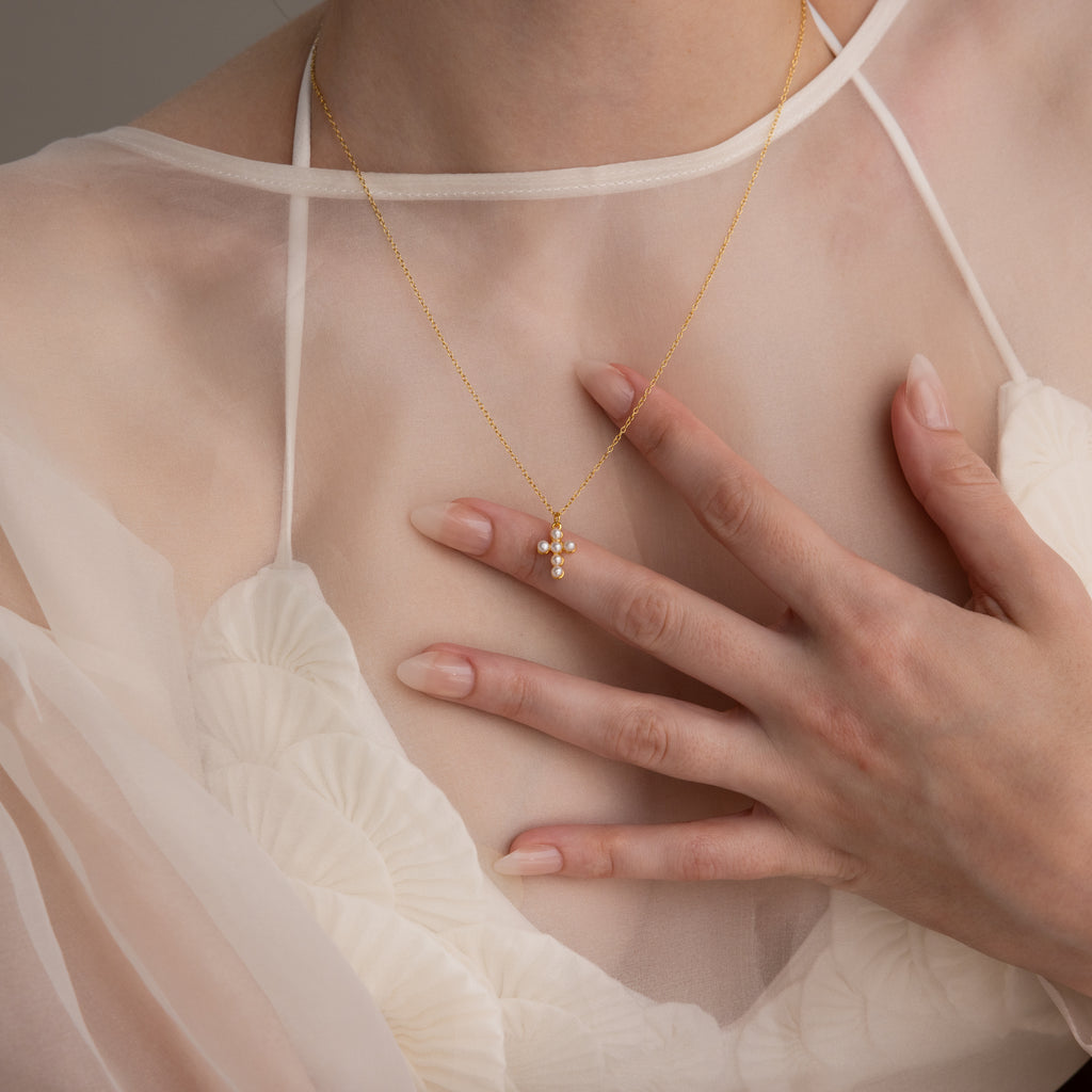 A woman in a sheer white dress touches her chest, highlighting the Pearl Cross Necklace in 18K Gold adorned with a luminous pearl cross charm.