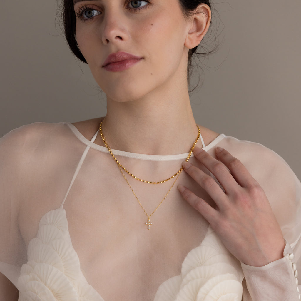 Woman wearing a sheer white top and layered gold necklaces, including the Pearl Cross Necklace featuring luminous pearls.