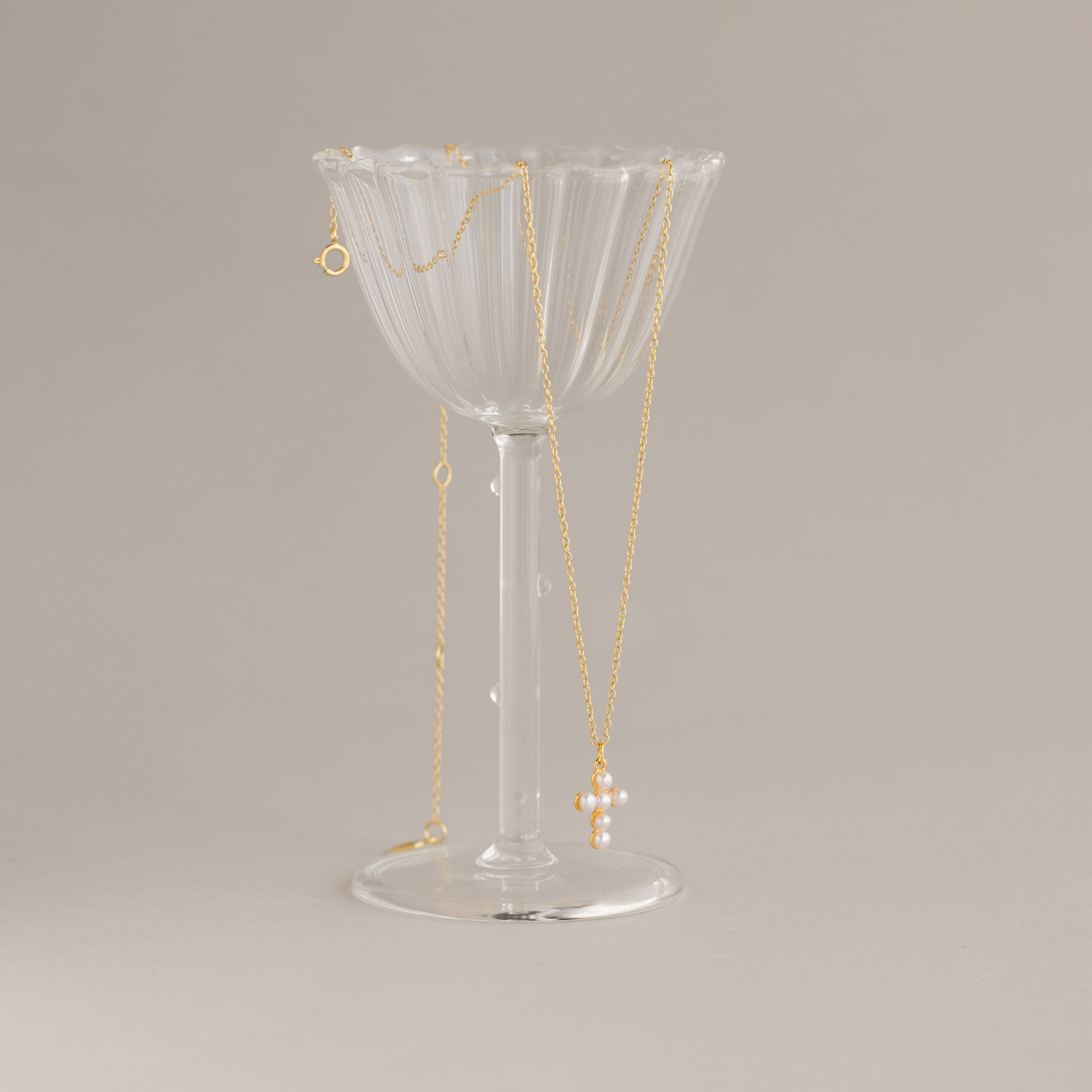 The Pearl Cross Necklace in 18K Gold, featuring luminous pearls and a cross charm, draped over a clear, ribbed glass cup on a neutral background.