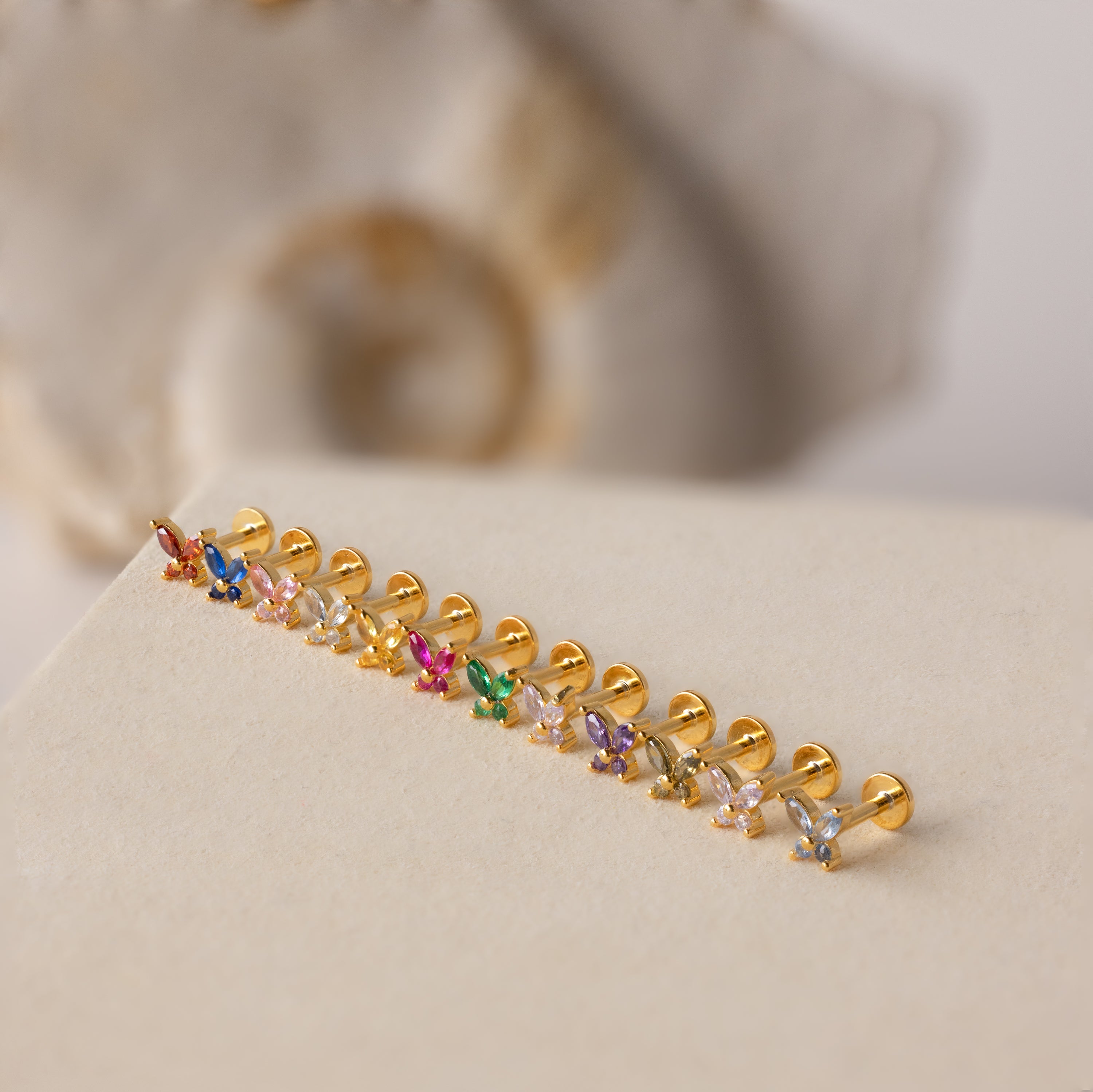 Butterfly Blue Zircon Flatback Studs with colorful designs are lined up on a beige surface against a blurred background.