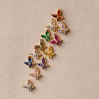 Eleven Butterfly Amethyst Flatback Studs with vibrant gemstones are arranged diagonally on a beige background, highlighting a unique take on personalized jewelry.