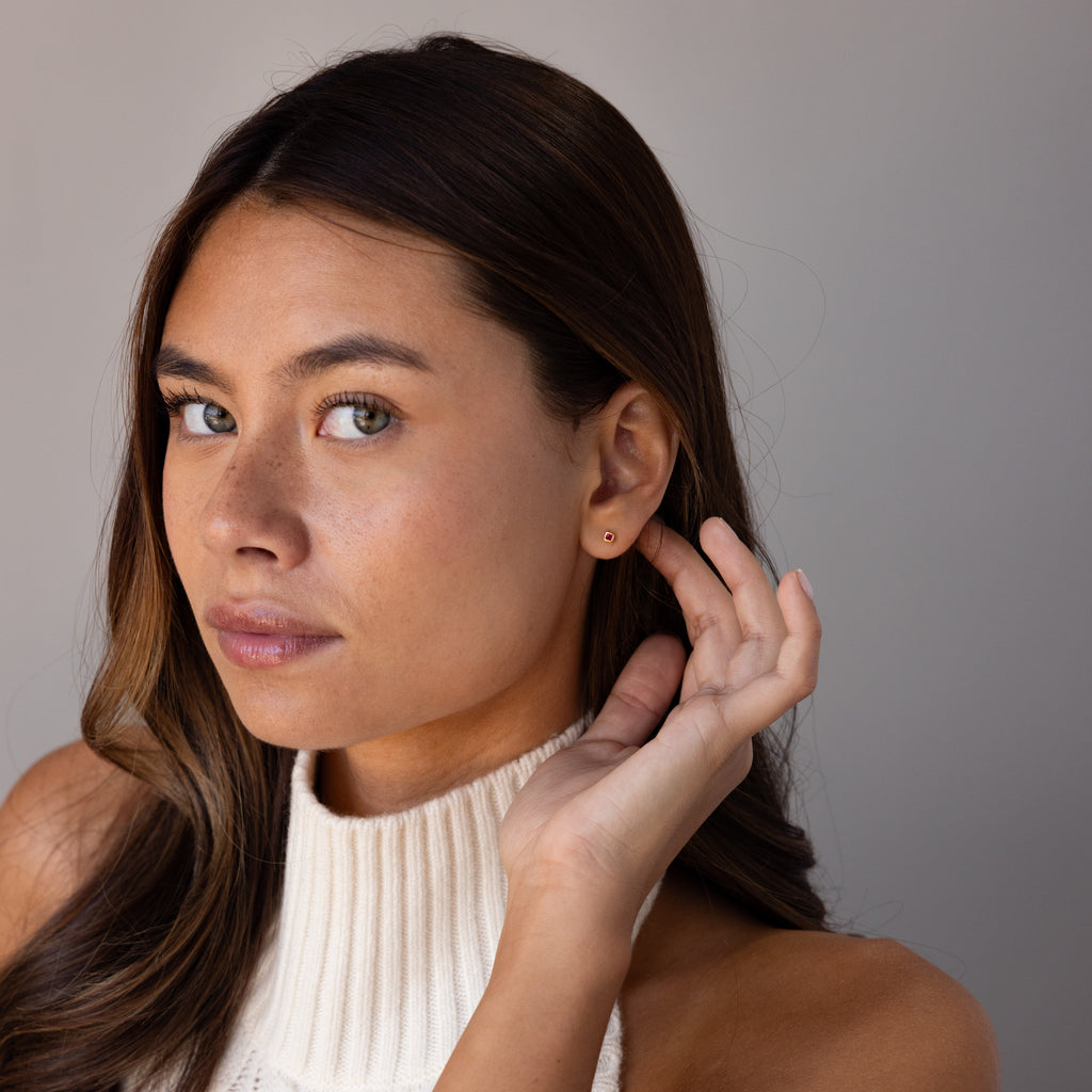 Woman with long brown hair in a sleeveless white top touches her ear and looks at the camera, highlighting Square Bezel Birthstone Flatback Studs.