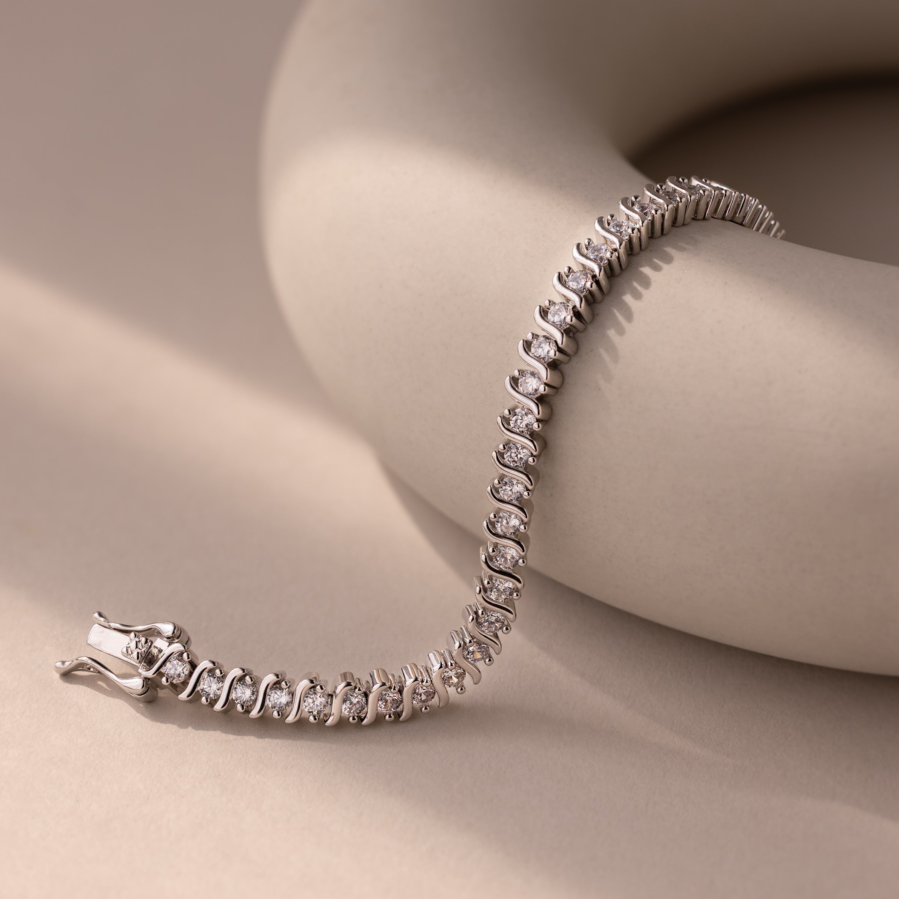 The S-Link Diamond Tennis Bracelet in White Gold is elegantly displayed on a smooth, curved beige surface in soft lighting, highlighting its timeless sophistication and classic appeal.