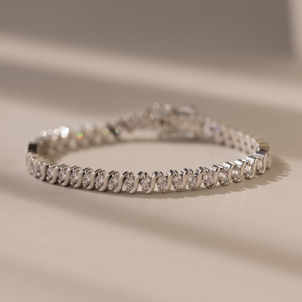 An elegant jewelry piece, the S-Link Diamond Tennis Bracelet in White Gold features round diamonds set in white gold, beautifully displayed on a beige surface under soft lighting.