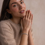 A woman in a beige sweater clasps her hands near her face, showcasing the elegant S-Link Diamond Tennis Bracelet in White Gold as she looks at the camera.