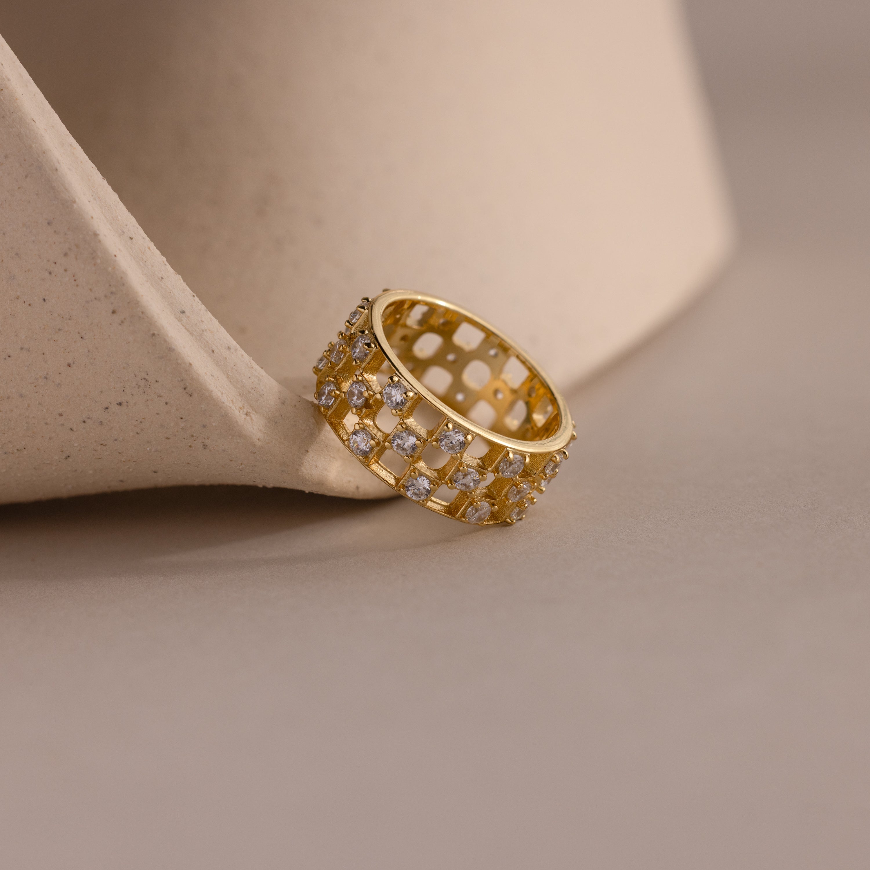 Close-up of a gold checkered diamond ring, featuring a wide band with open grid design and sparkling round-cut stones, resting against a ceramic surface.