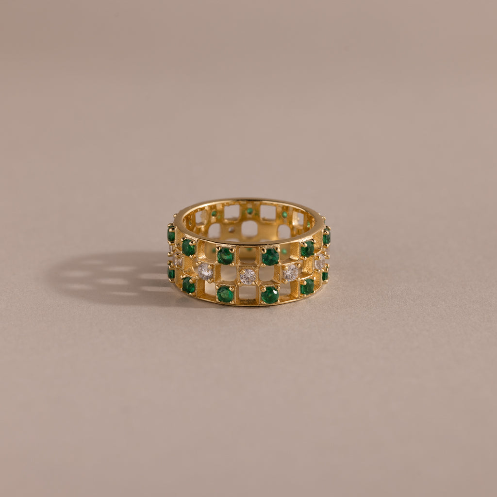 Gold checkered band ring featuring alternating rows of emerald and diamond round-cut CZ stones set in an openwork grid design, displayed on a neutral background.