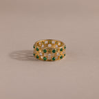 Gold checkered band ring featuring alternating rows of emerald and diamond round-cut CZ stones set in an openwork grid design, displayed on a neutral background.