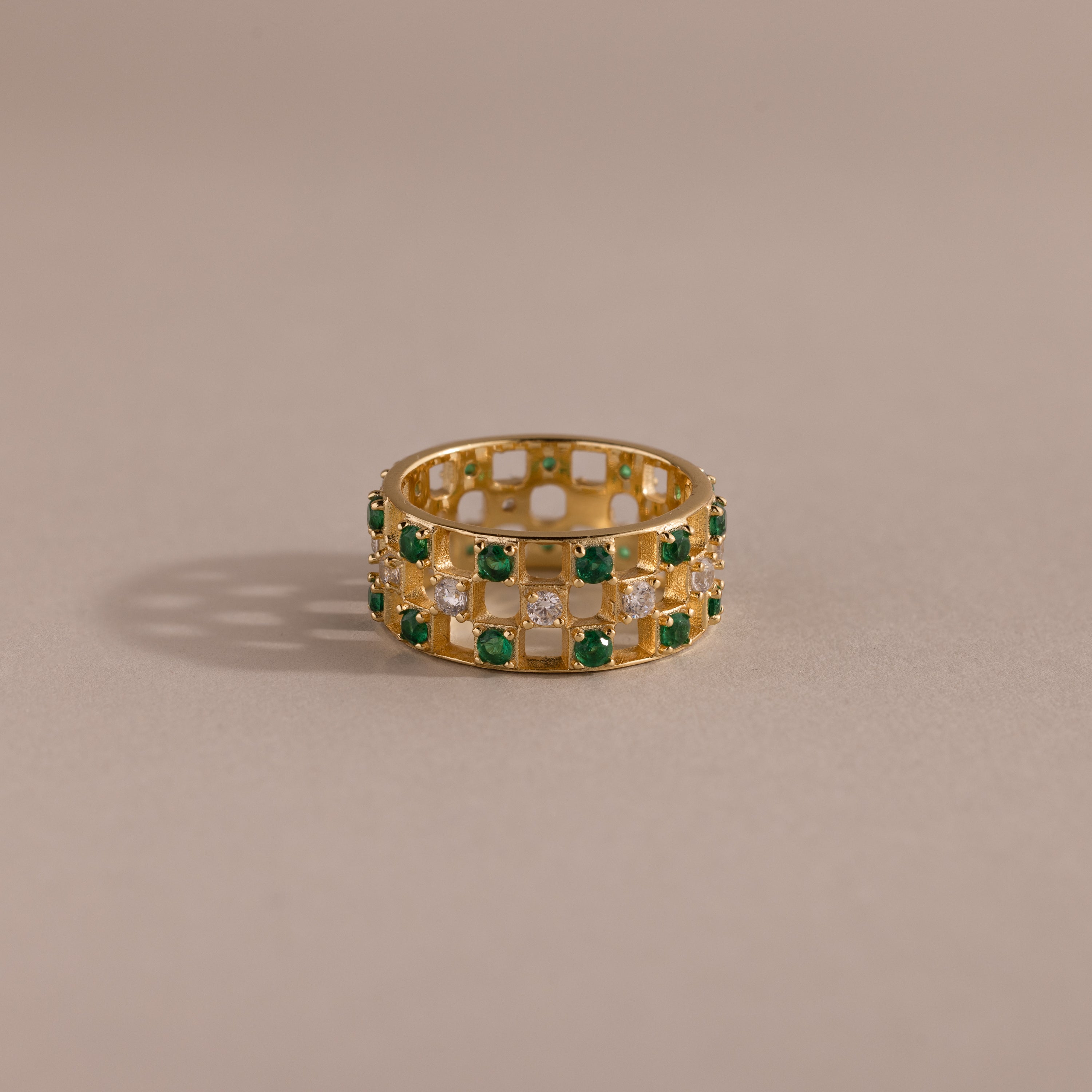 Gold checkered band ring featuring alternating rows of emerald and diamond round-cut CZ stones set in an openwork grid design, displayed on a neutral background.