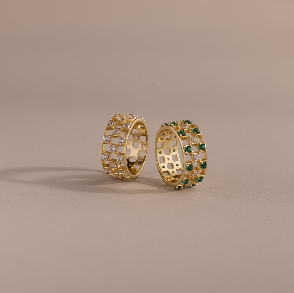 Two checkered band rings, one with diamonds and one with emeralds, each round-cut CZ stones set in an openwork grid design, displayed on a neutral background.