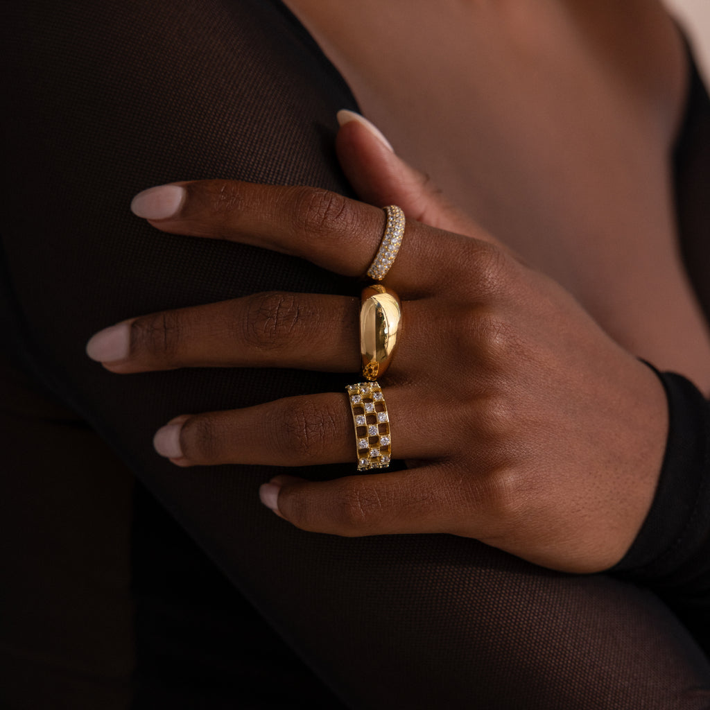 Close-up of model wearing a gold checkered diamond ring with an open grid design and sparkling round-cut stones, stacked with matching gold rings.