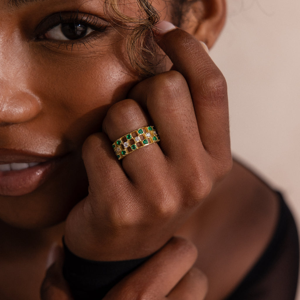 Model wearing a gold checkered band ring featuring alternating rows of emerald and diamond round-cut CZ stones set in an openwork grid design.
