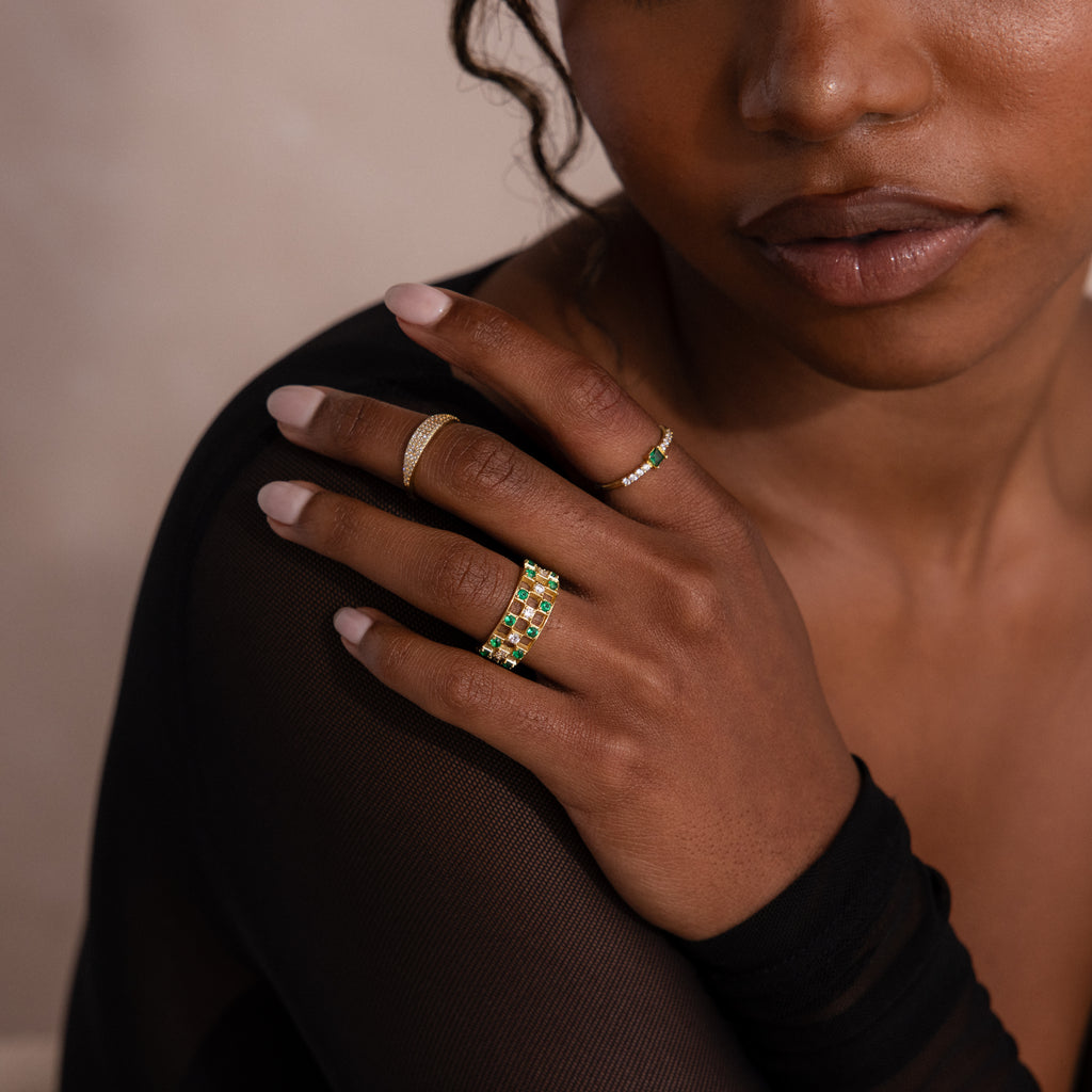 Model wearing gold checkered band ring featuring alternating rows of emerald and diamond round-cut CZ stones set in an openwork grid design, stacked with multiple gold rings.