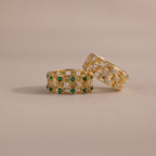 Two checkered band rings, one with diamonds and one with emeralds, each round-cut CZ stones set in an openwork grid design, displayed on a neutral background.