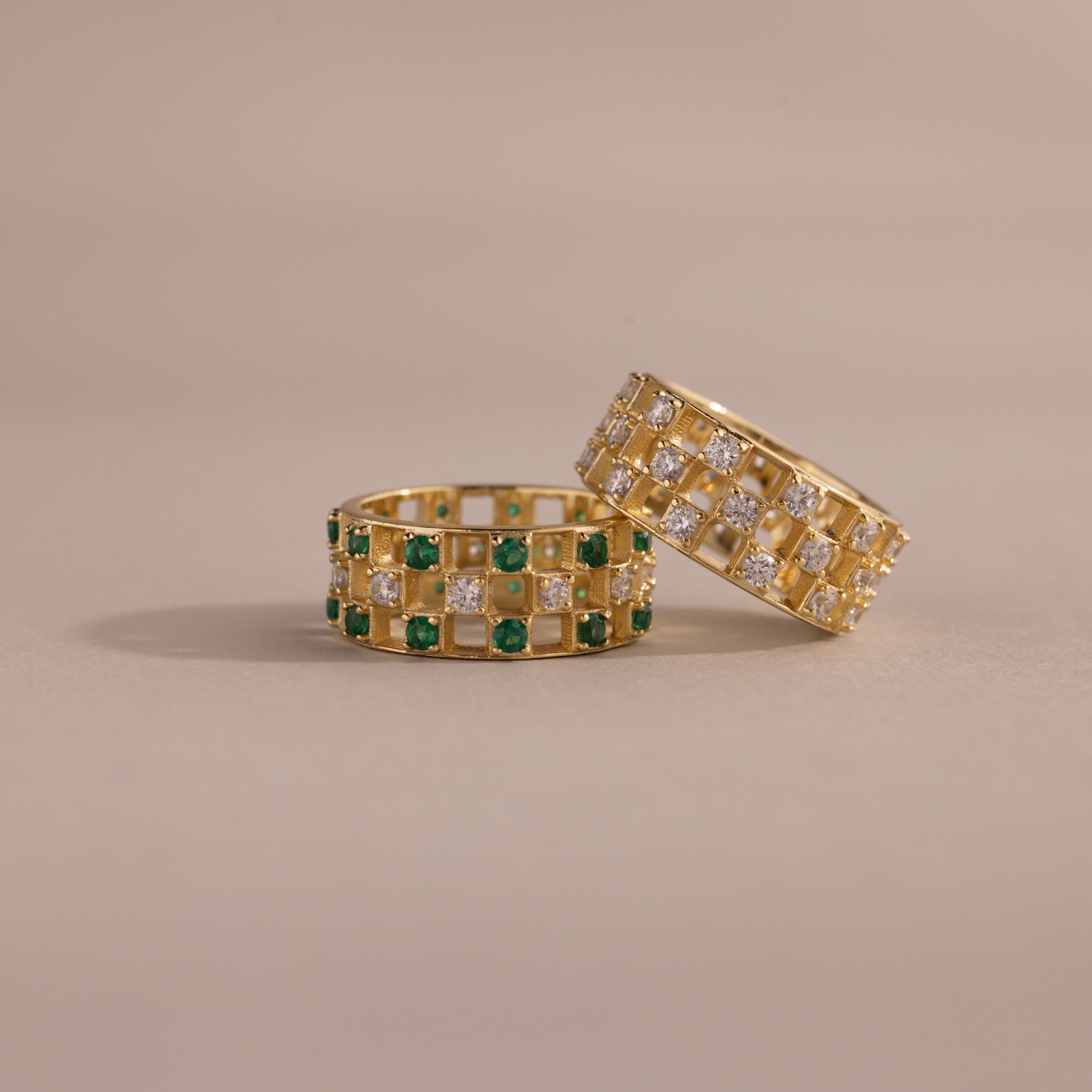 Two checkered band rings, one with diamonds and one with emeralds, each round-cut CZ stones set in an openwork grid design, displayed on a neutral background.