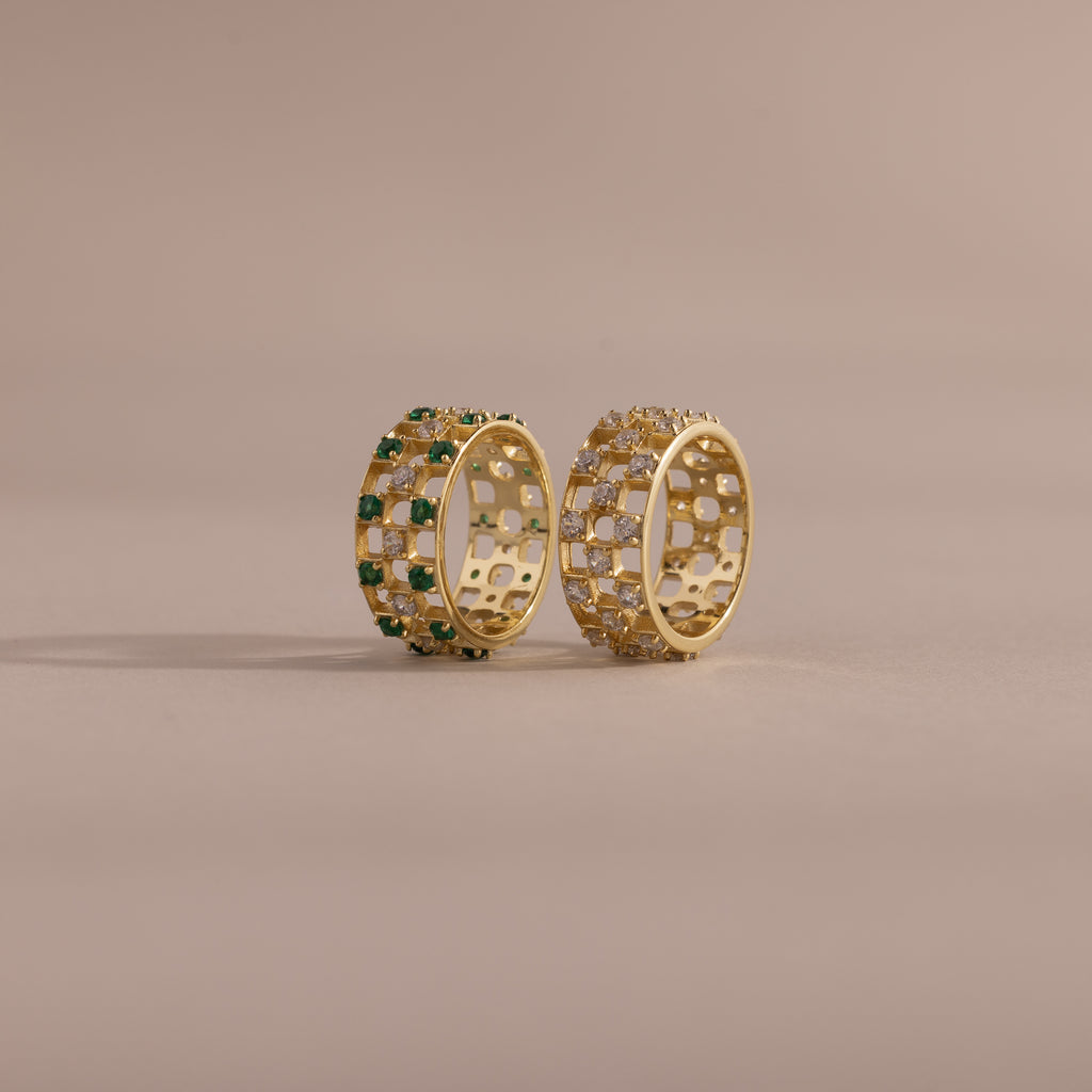 Two checkered band rings, one with diamonds and one with emeralds, each round-cut CZ stones set in an openwork grid design, displayed on a neutral background.