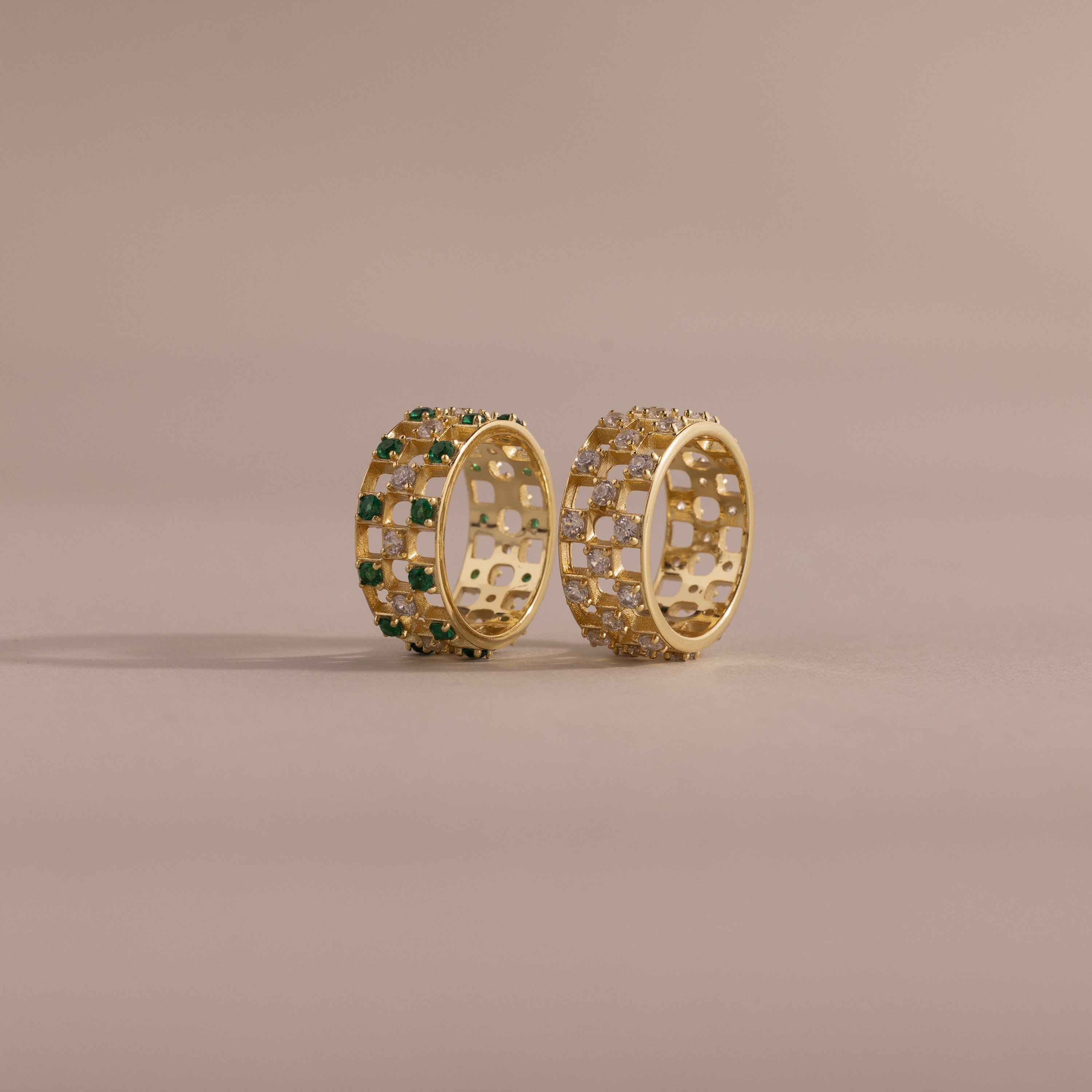 Two checkered band rings, one with diamonds and one with emeralds, each round-cut CZ stones set in an openwork grid design, displayed on a neutral background.