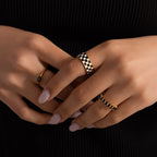 Model wearing a thick gold band ring with a black and white enamel checkerboard pattern, stacking with multiple gold rings with black onyx gemstones.