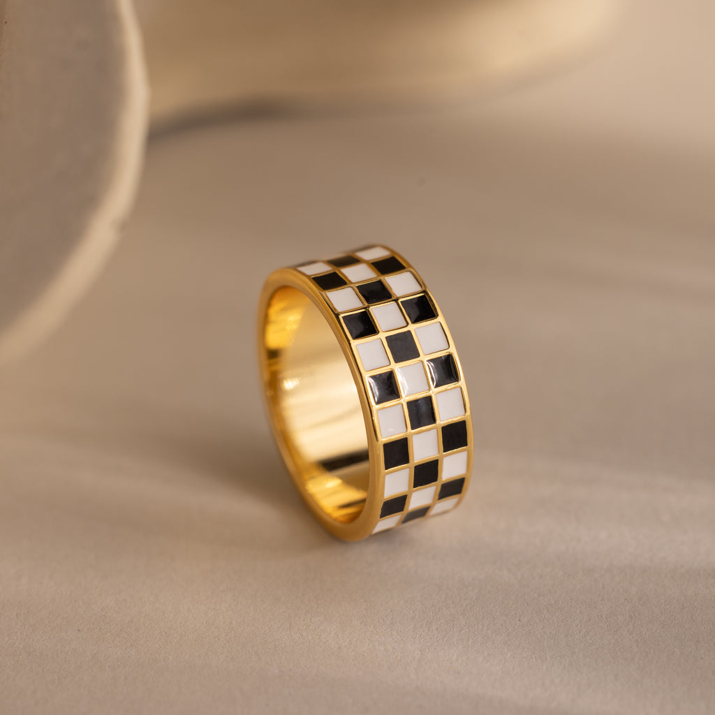 Gold ring with a thick band featuring a black and white enamel checkerboard pattern positioned up-right on a neutral background.