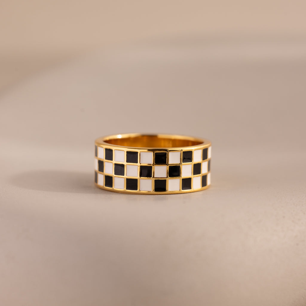 Front-view of a gold ring with a thick band featuring a black and white enamel checkerboard pattern resting on a neutral surface.