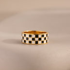 Front-view of a gold ring with a thick band featuring a black and white enamel checkerboard pattern resting on a neutral surface.