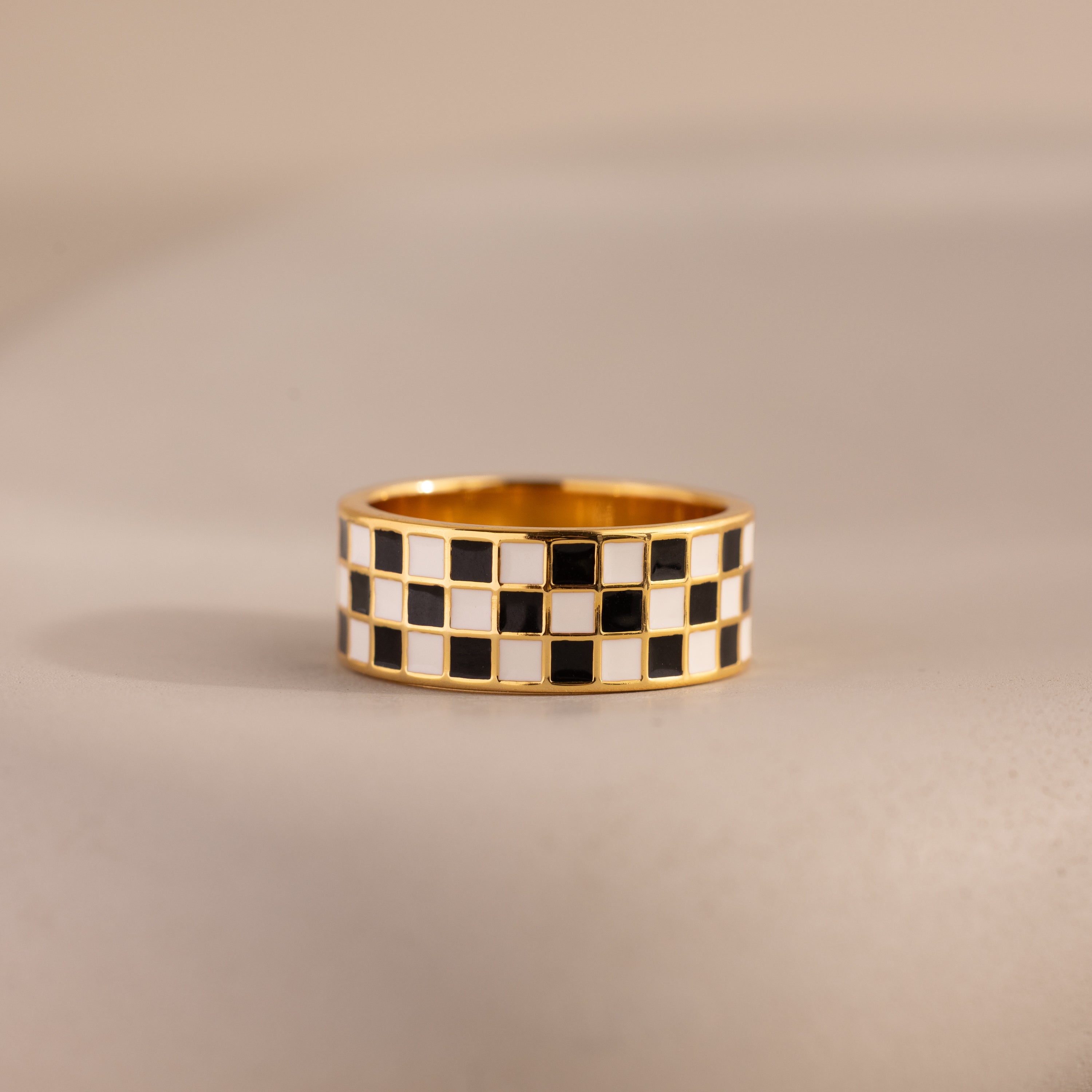 Front-view of a gold ring with a thick band featuring a black and white enamel checkerboard pattern resting on a neutral surface.