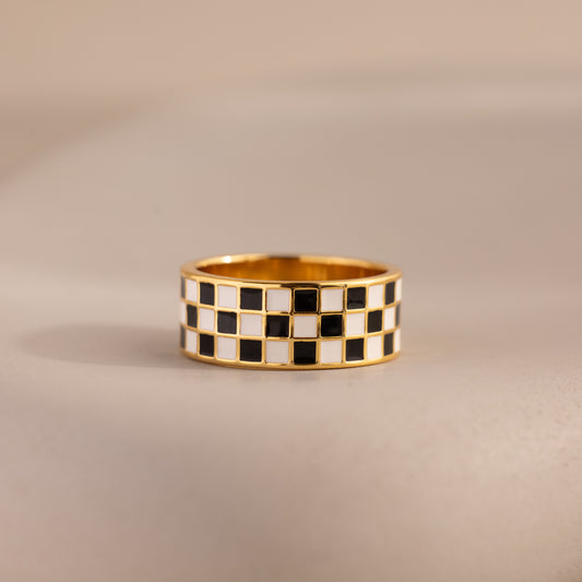 Front-view of a gold ring with a thick band featuring a black and white enamel checkerboard pattern resting on a neutral surface.