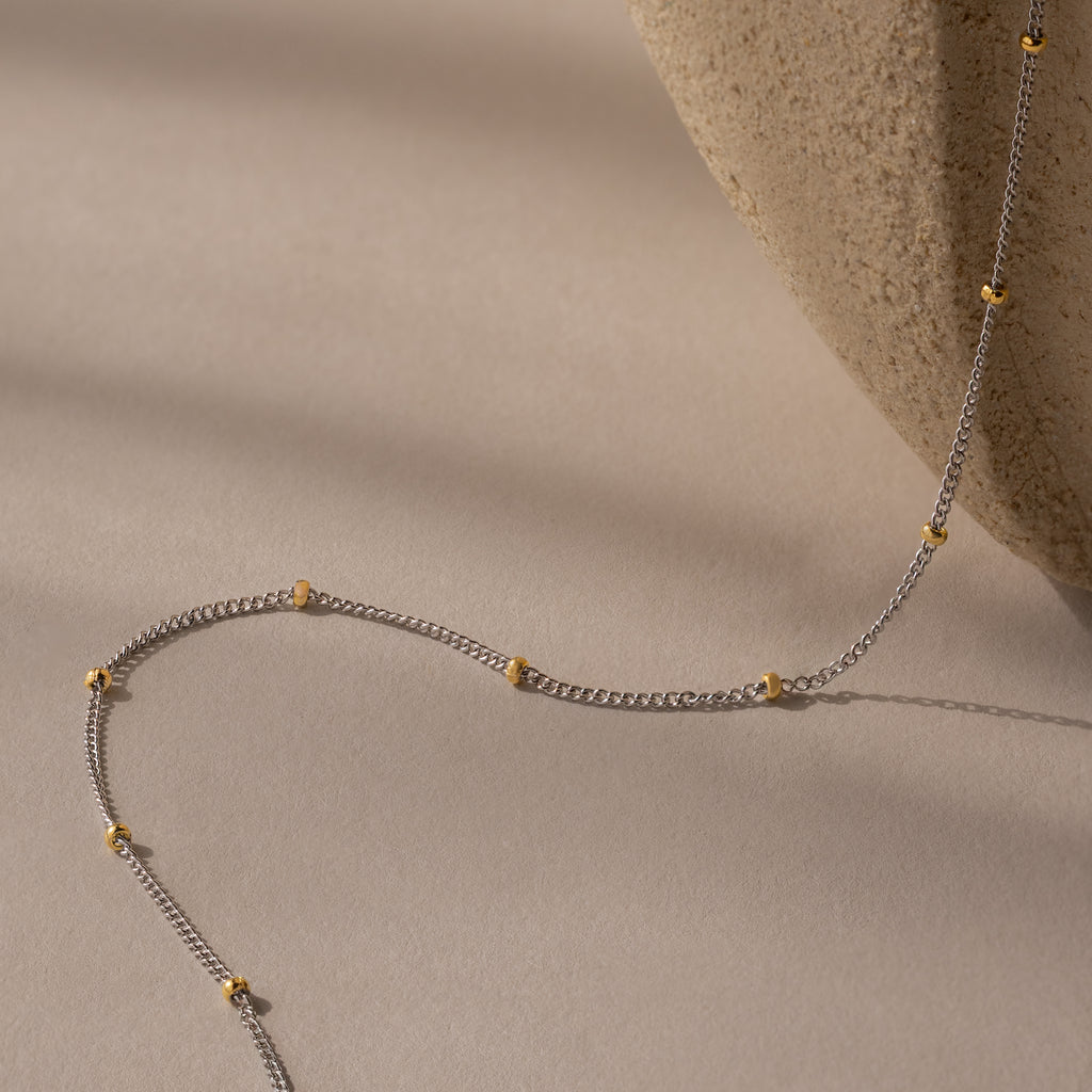 The Mixed Metal Beaded Necklace features a silver satellite chain with gold beads, elegantly displayed on beige beside a textured stone. Ideal for layering, it's perfect for any occasion.