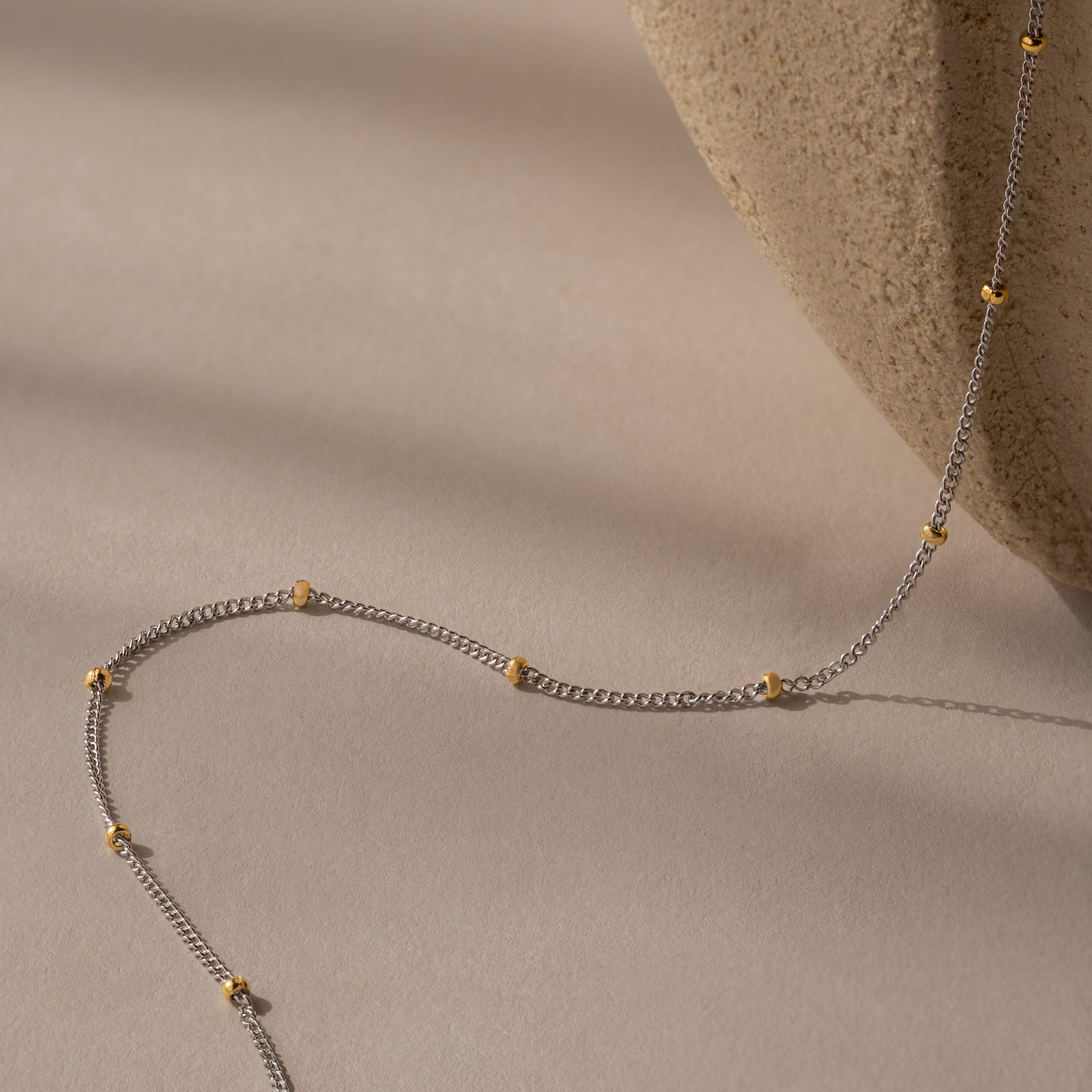 The Mixed Metal Beaded Necklace features a silver satellite chain with gold beads, elegantly displayed on beige beside a textured stone. Ideal for layering, it's perfect for any occasion.