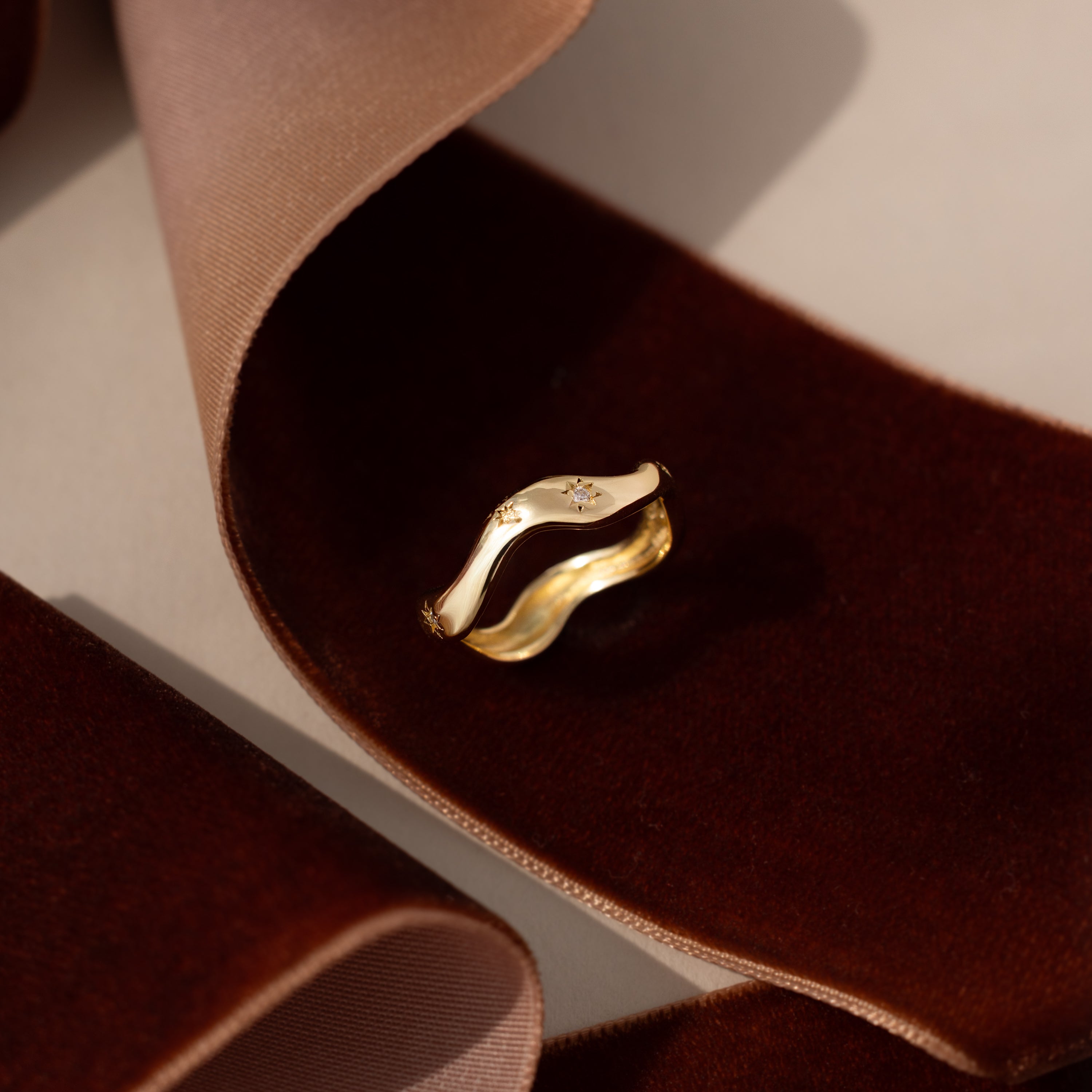 The Wavy Diamond Celestial Ring, featuring a sparkling diamond accent, is elegantly displayed on soft brown velvet ribbons.