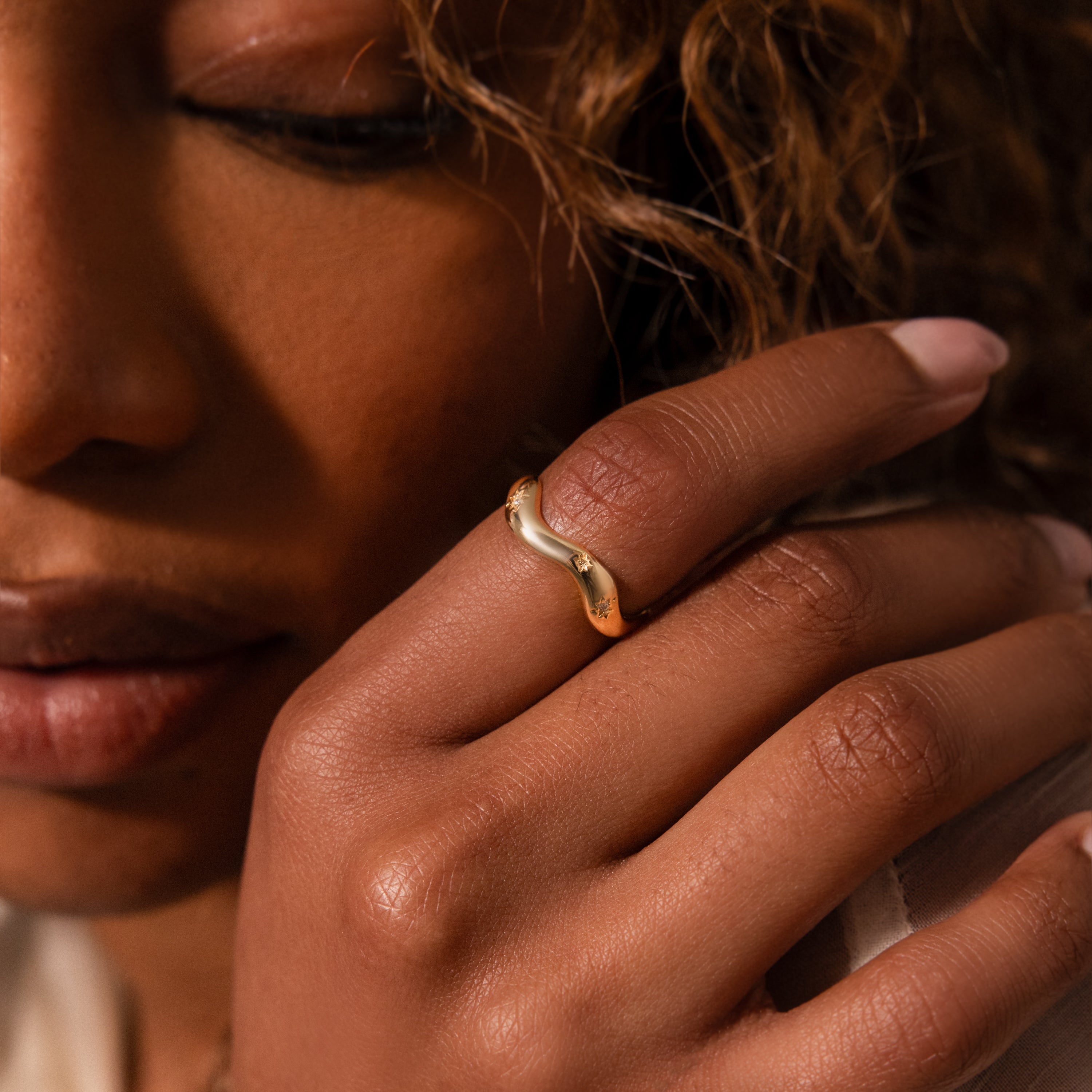 A hand wearing the Wavy Diamond Celestial Ring is shown close to a woman's face with curly hair.