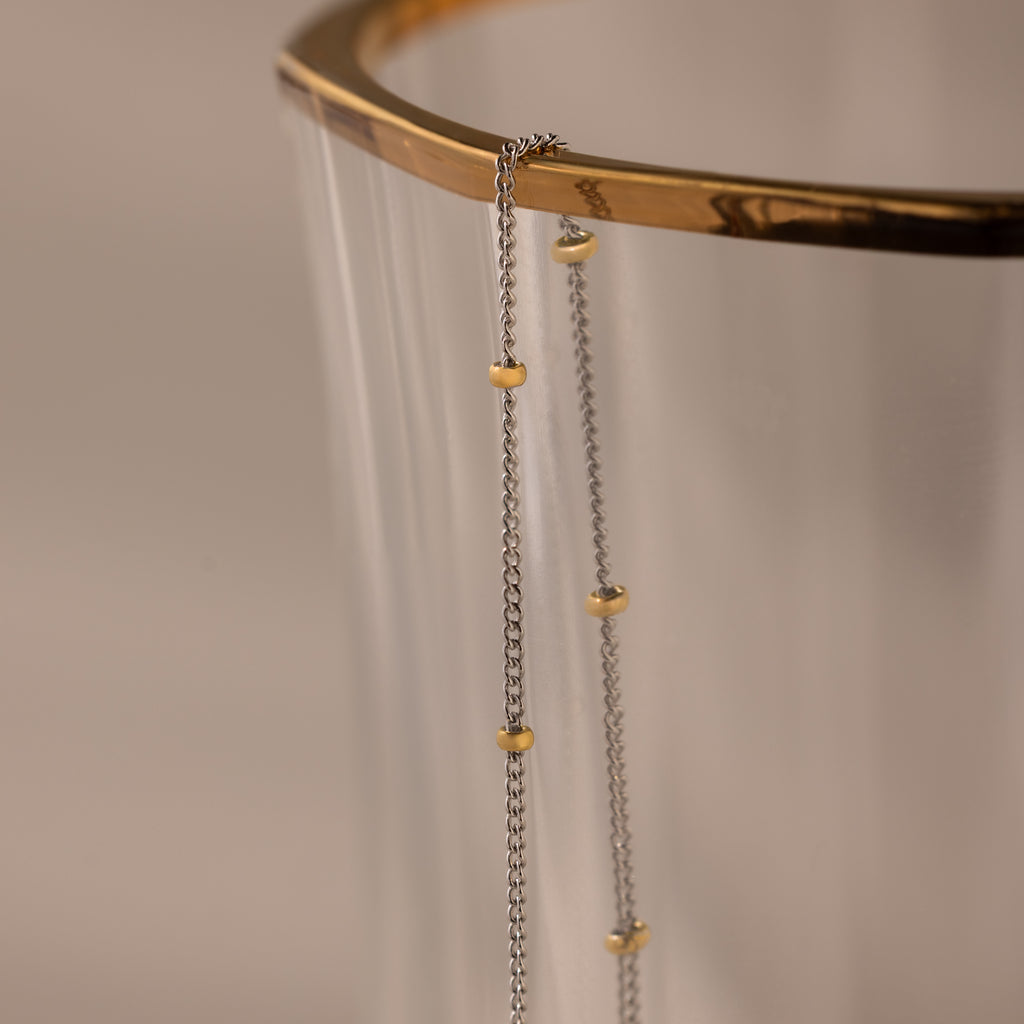 The Mixed Metal Beaded Necklace features a silver satellite chain with small gold beads, making it perfect for layering and adding an elegant touch to any outfit.