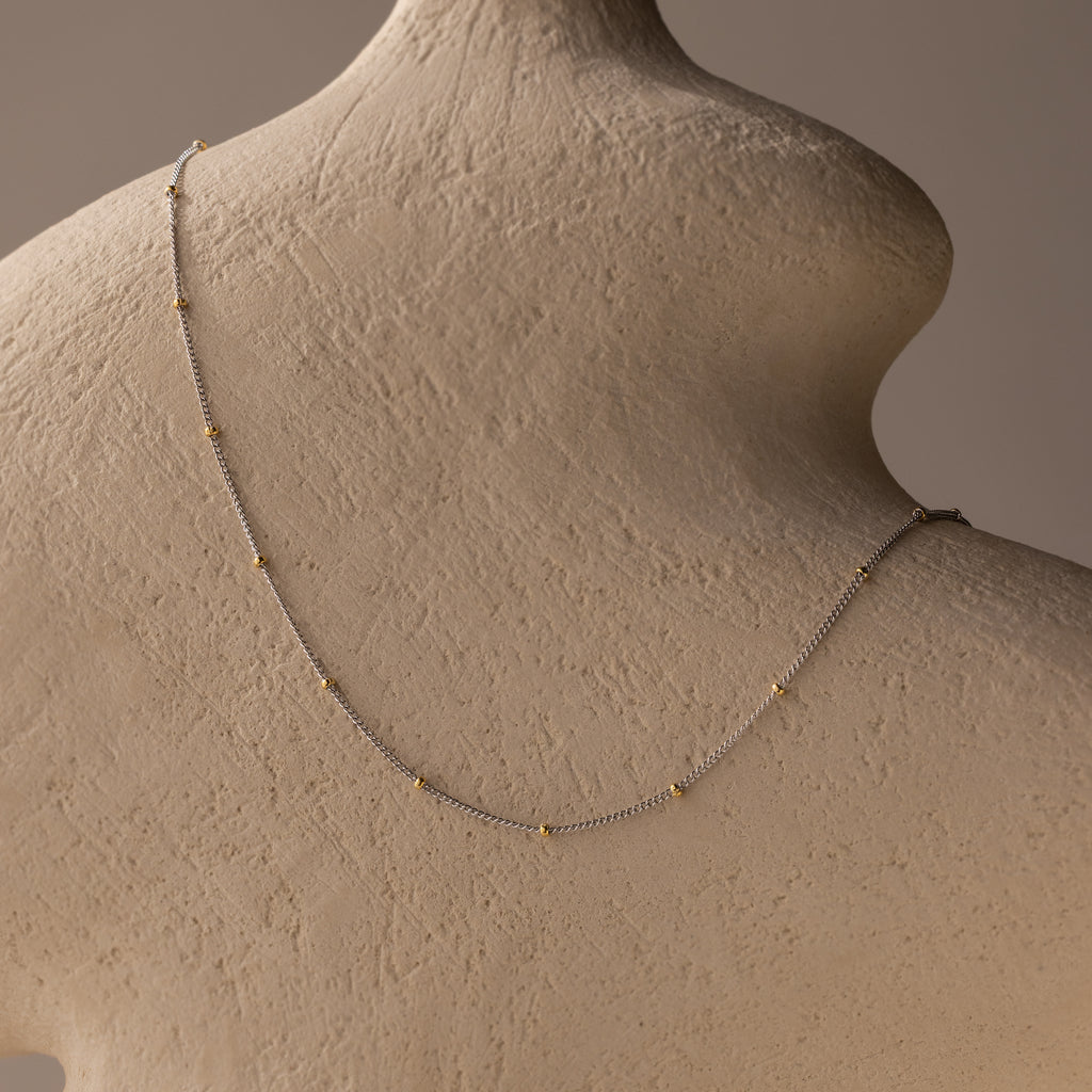 The Mixed Metal Beaded Necklace features delicate beads on a satellite chain, showcased draped across the back of a textured beige mannequin bust.